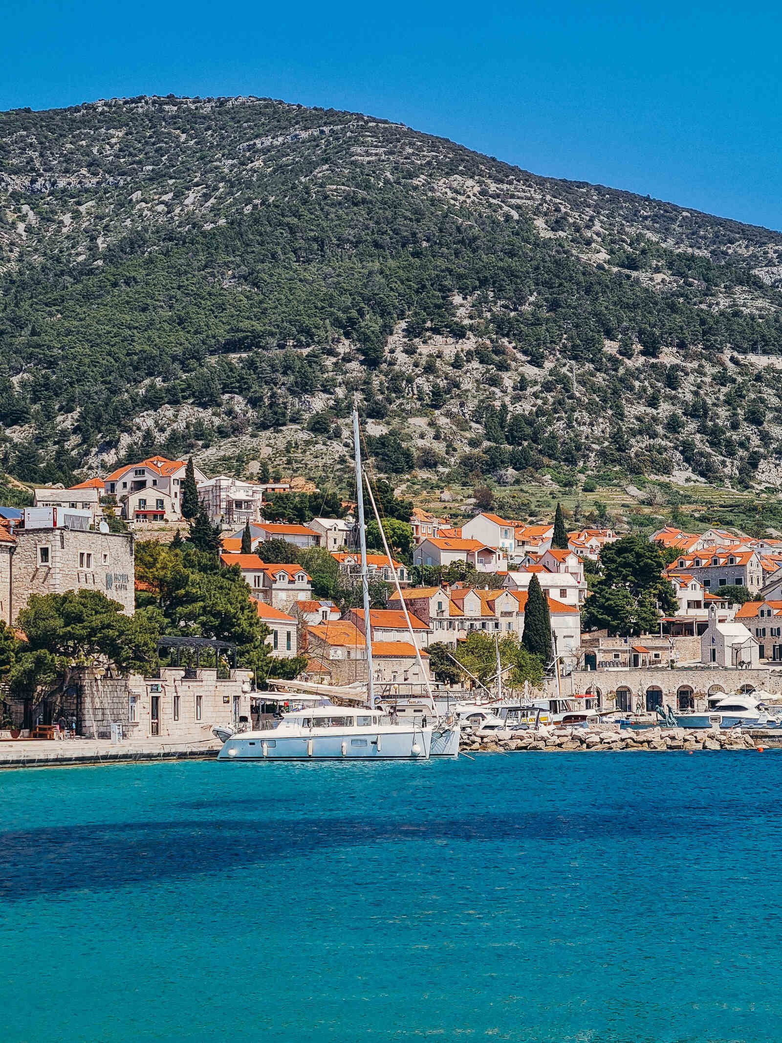 Looking across blue sea to a town on the hillside with houses made of stone and orange rooftops. A catamaran is docked in front of the town. A mountain towers behind the town