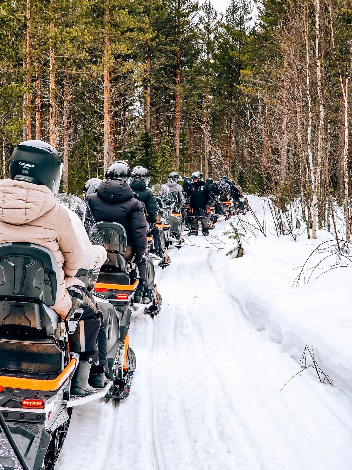 Many snowmobiles lined up along a snowy trail waiting to go