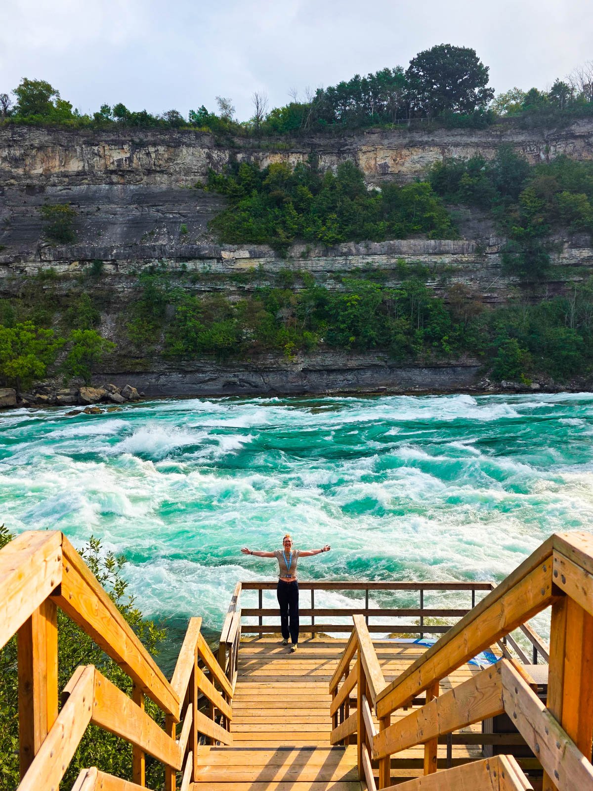 a wooden staircase down to a rapid flowing river with turquoise water and white rapids flowing, a cliff face on the opposite side of the river