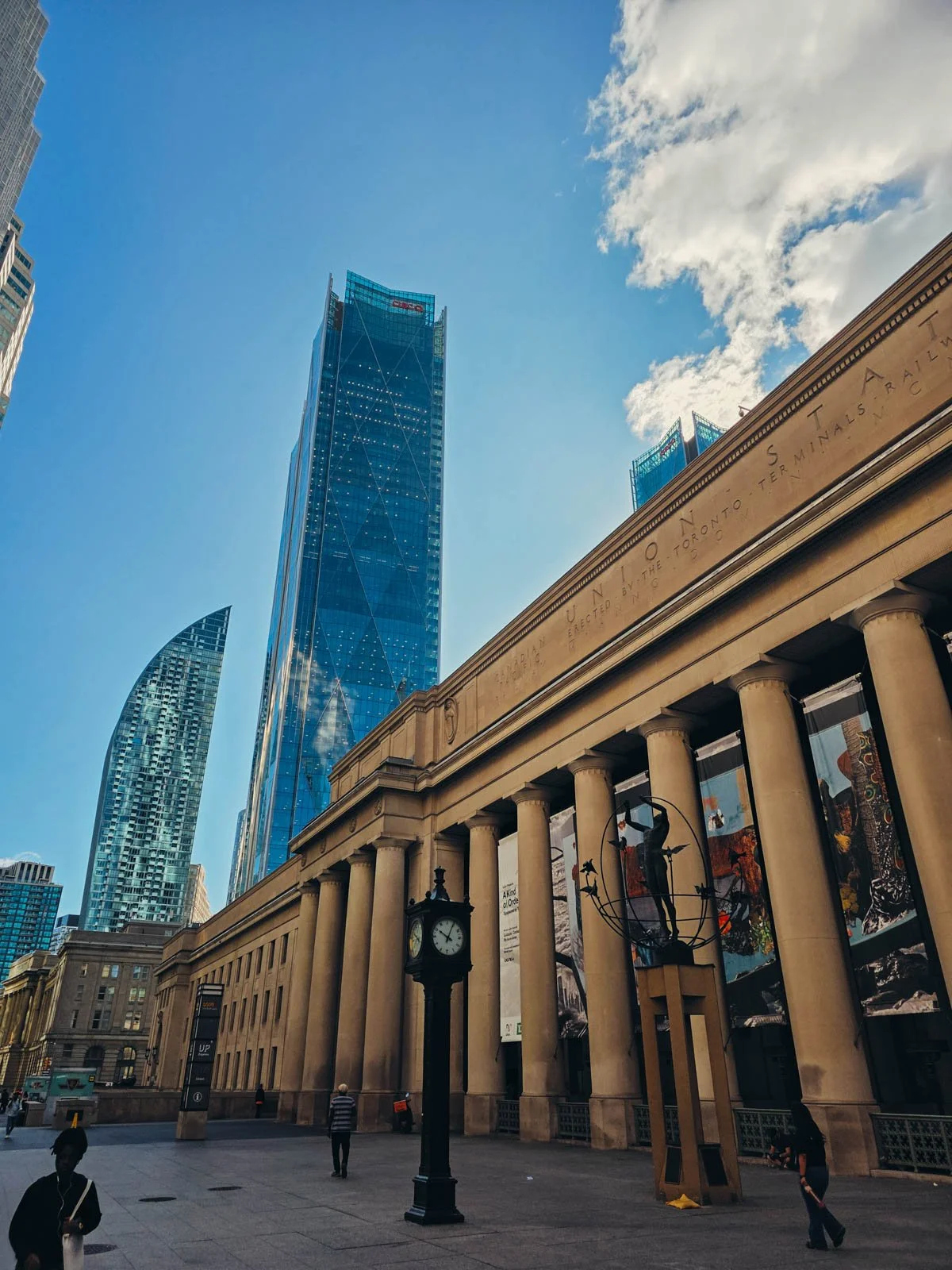 The large sandstone exterior of a train station with many large columns. Tall glass skyscrapers can be seen behind