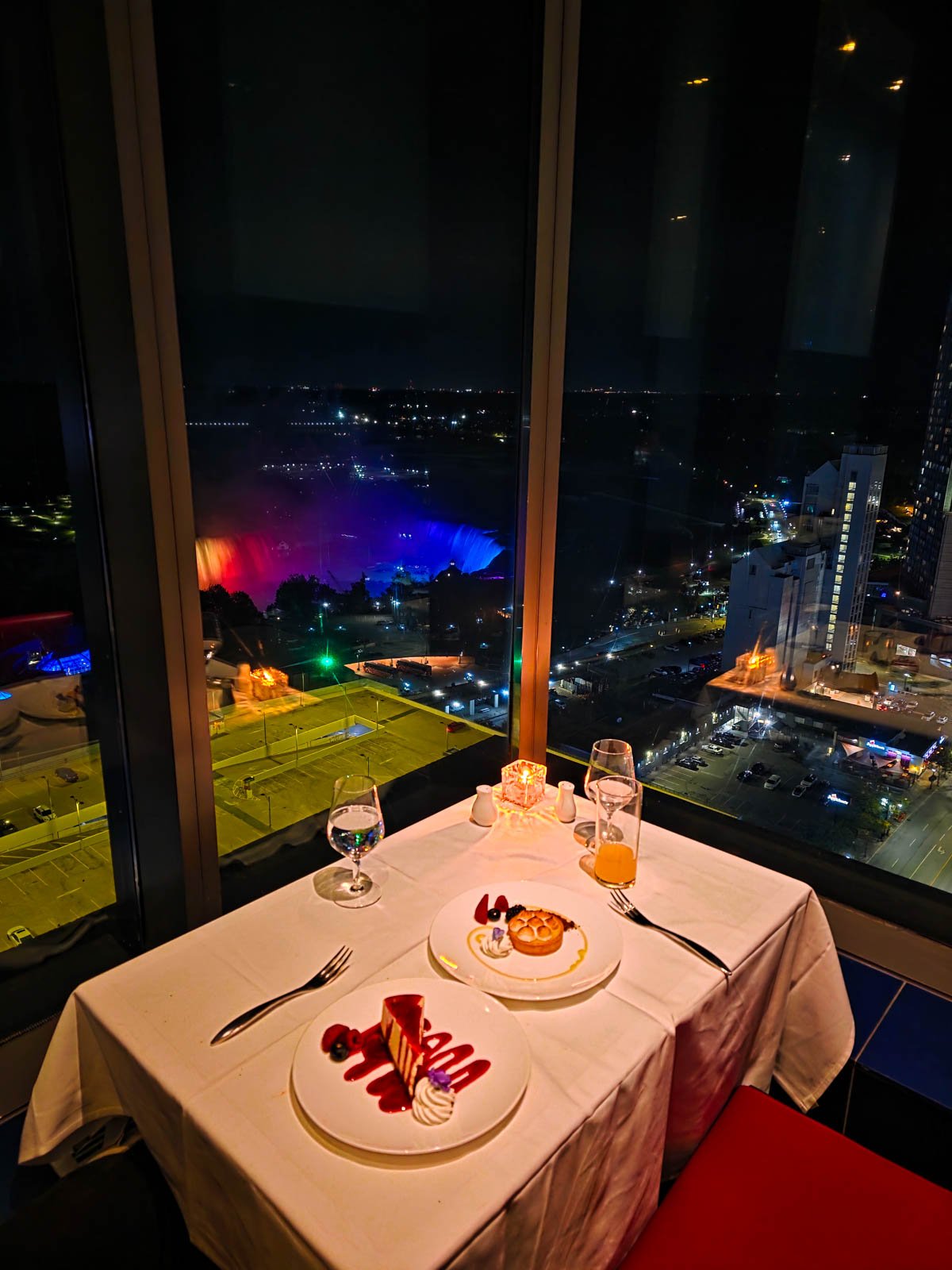 A white table covered in a tablecloth set in a corner of a window, with two plates of desserts next to a large window with a view of Niagara Falls lit up at night