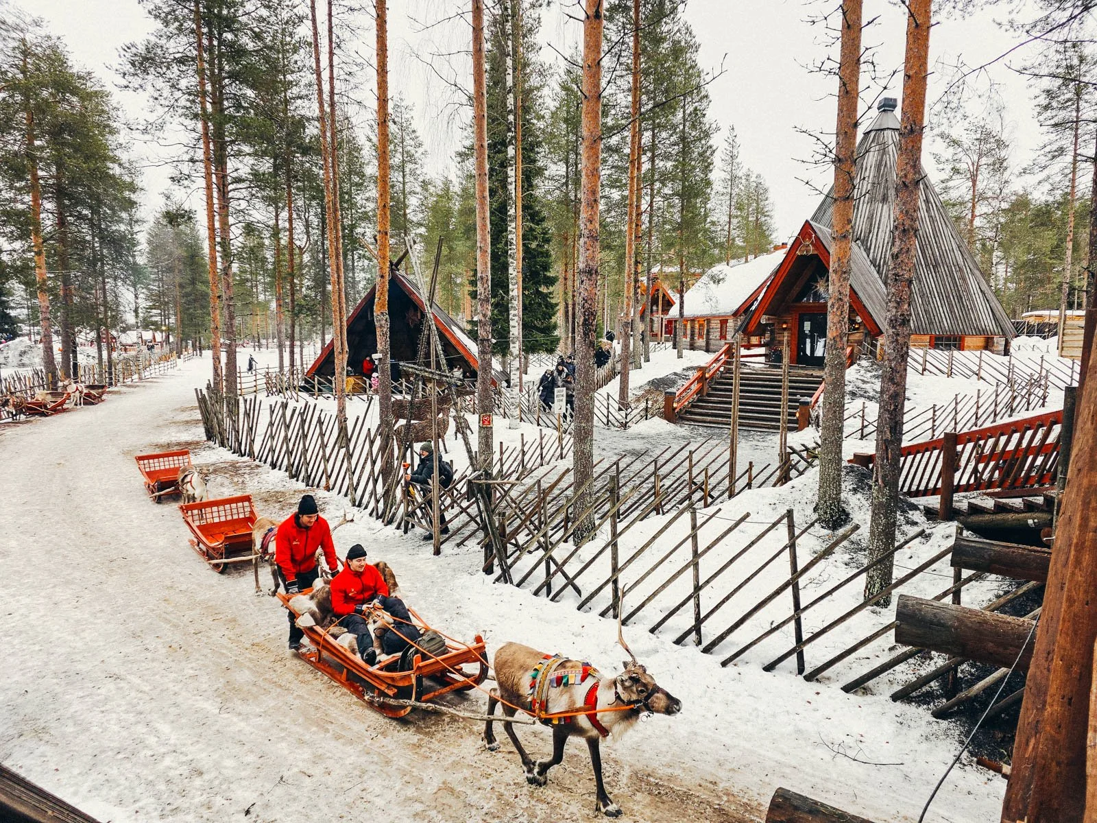 Two people in red being pulled in a sleigh down a path by reindeer in the snow. Two large pointed wooden building are in the background at Santa Claus Village - one of the best places to visit in Rovaniemi
