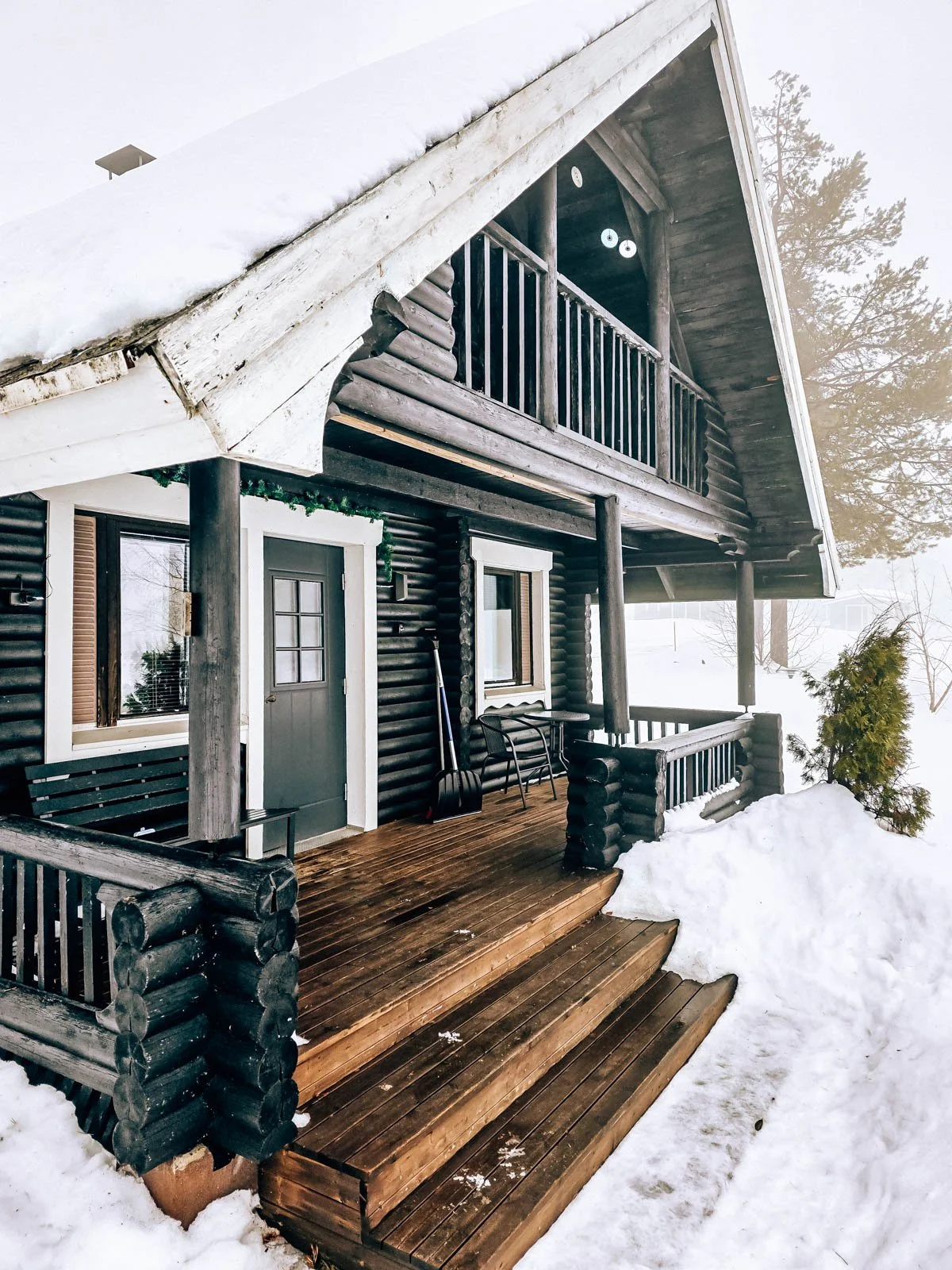 A dark coloured log cabin with a large deck surrounded by snow