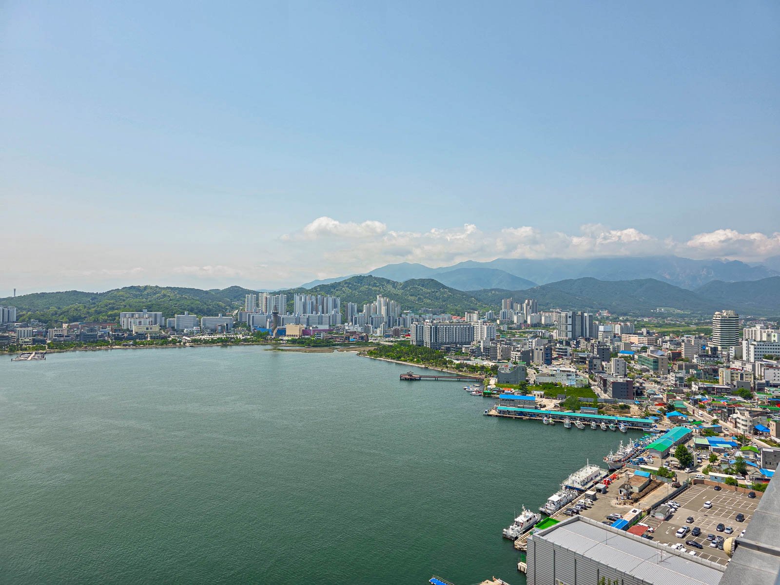 A view from a high up hotel room overlooking a lake, to the left on the lake shore is a city skyline with tall buildings and smaller buildings along the shore, behind the city are rolling green hills and mountains. It's a sunny clear day
