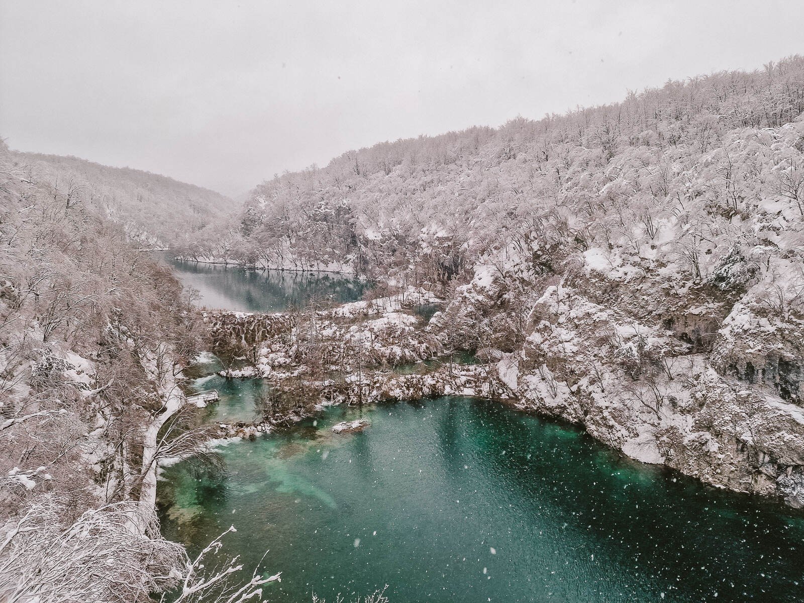Snow is falling on aview of a turquoise blue green lake in a canyon with snow covered cliffs and trees on either side and grey sky in Plitvice snow