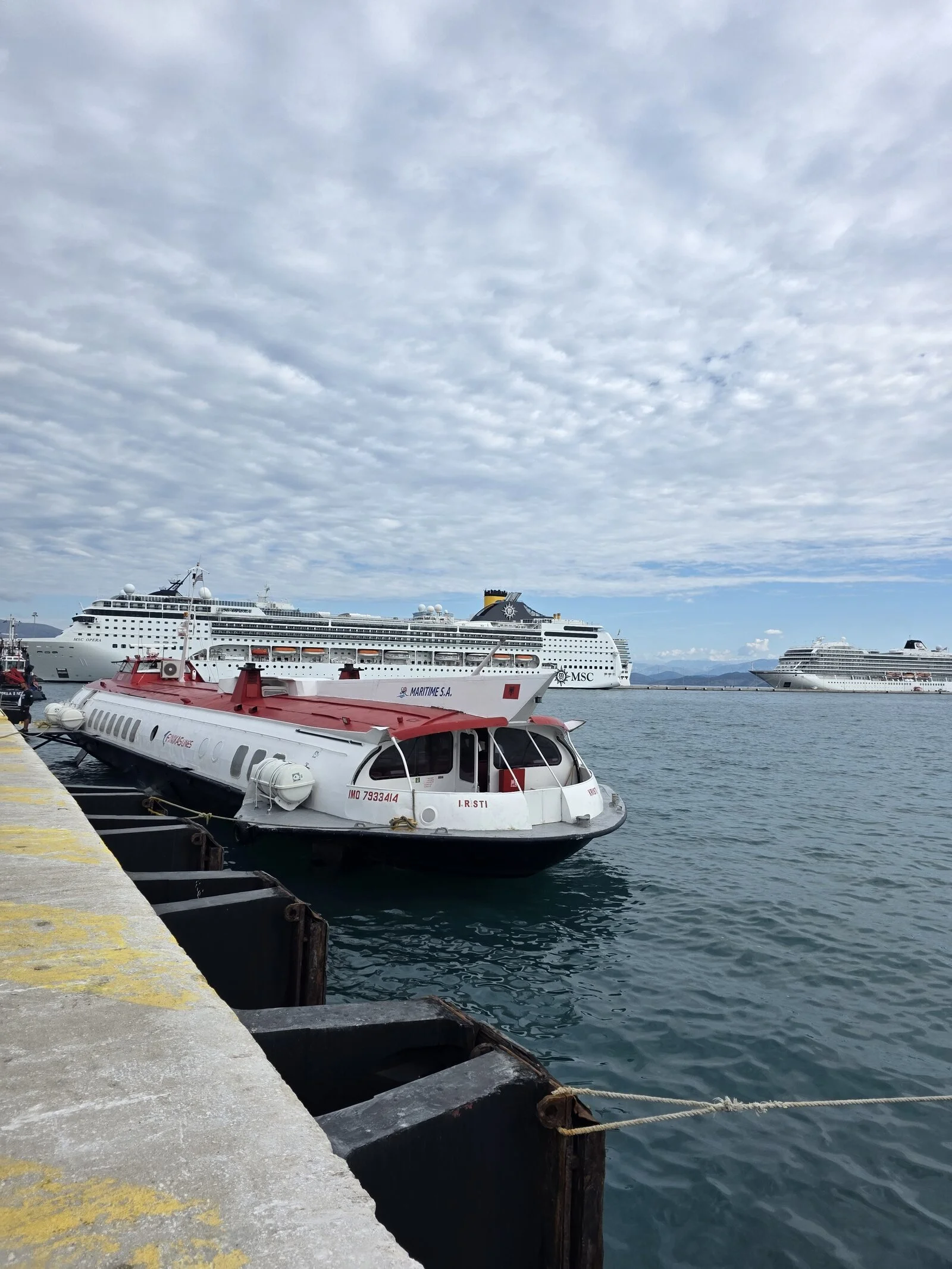 A large long hydrofoil ferry from Corfu to Albania docked along a concrete dock with two large cruise ships in the distance
