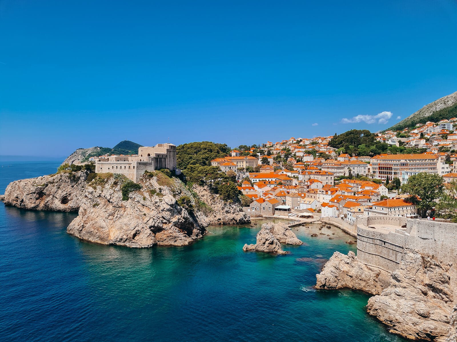 Looking across the coastline of Dubrovnik, a rocky cove can be seen below, stone houses with orange roofs and a large fortress on the top of the cliffs opposite