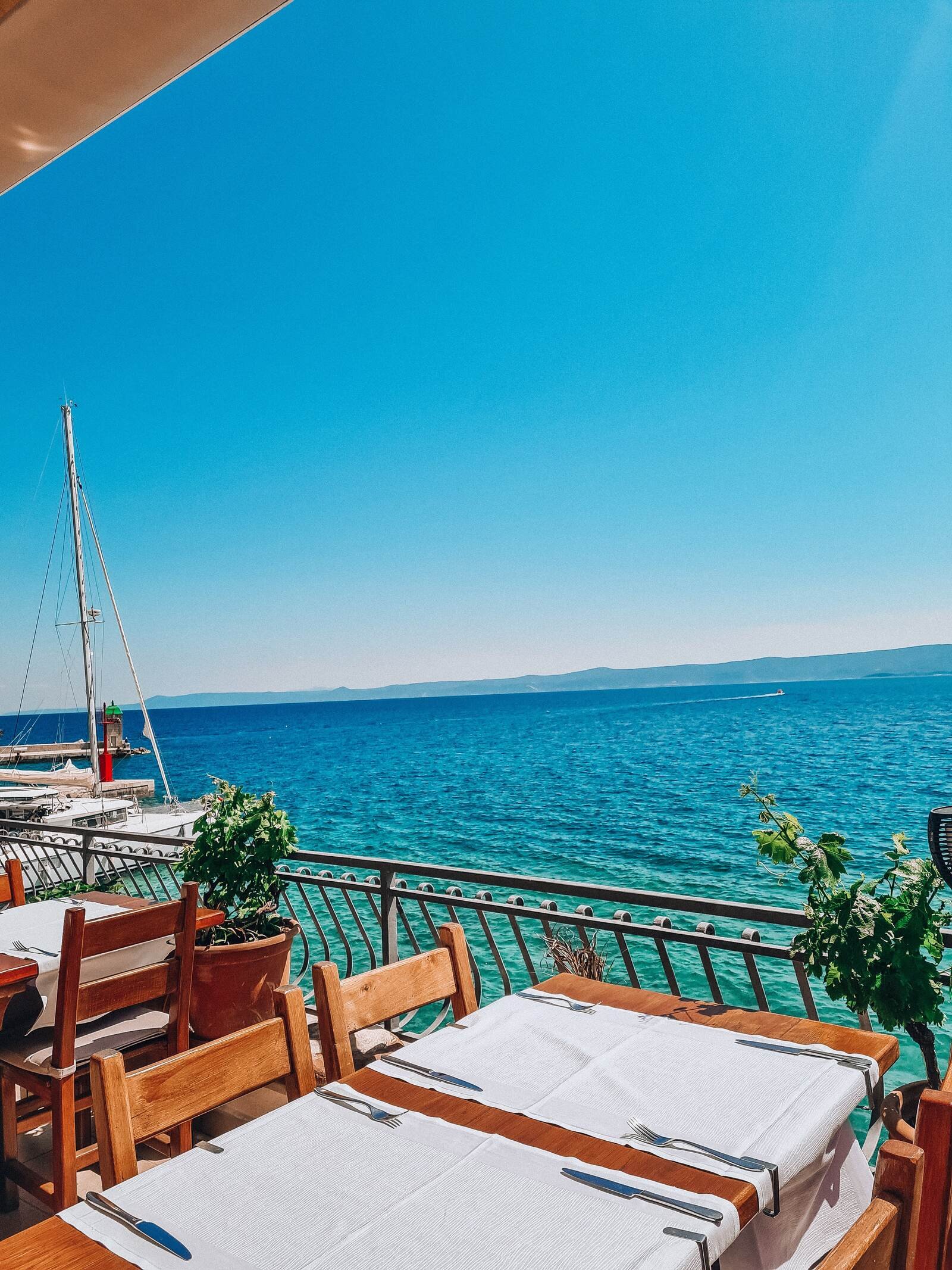 A wooden table on a sun terrace laid ready for people to eat. The table is right next to the terrace balcony and looks  out across blue water with a catamaran in it