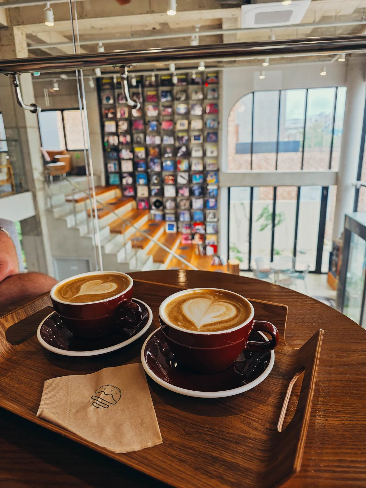 Two coffees on a wooden table, out of focus behind is a set of stairs next to a wall of colourful records