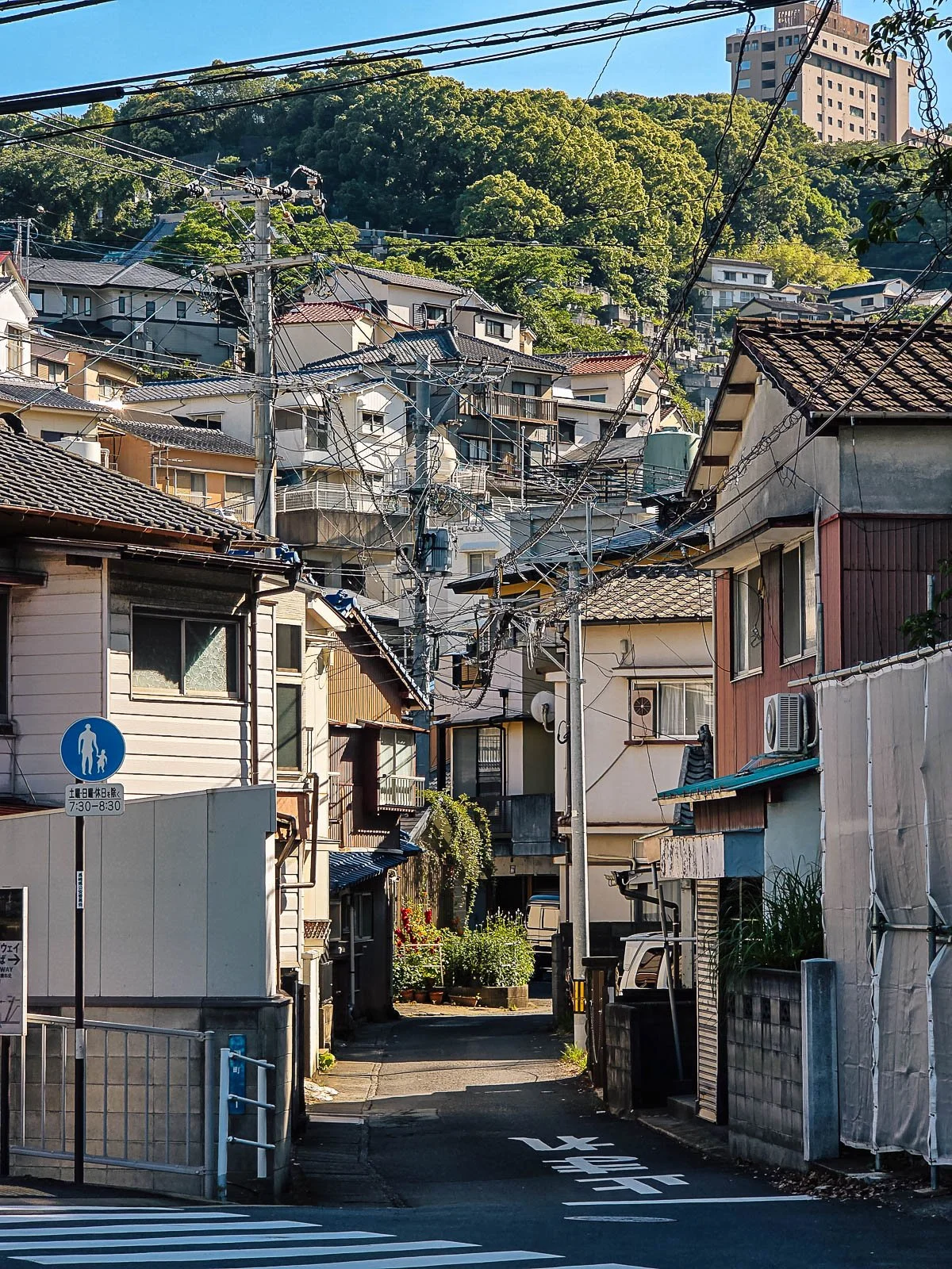 A narrow street in Nagasaki lined with Japanese houses, power lines are strung across the street and there is a hill covered with green trees in the background