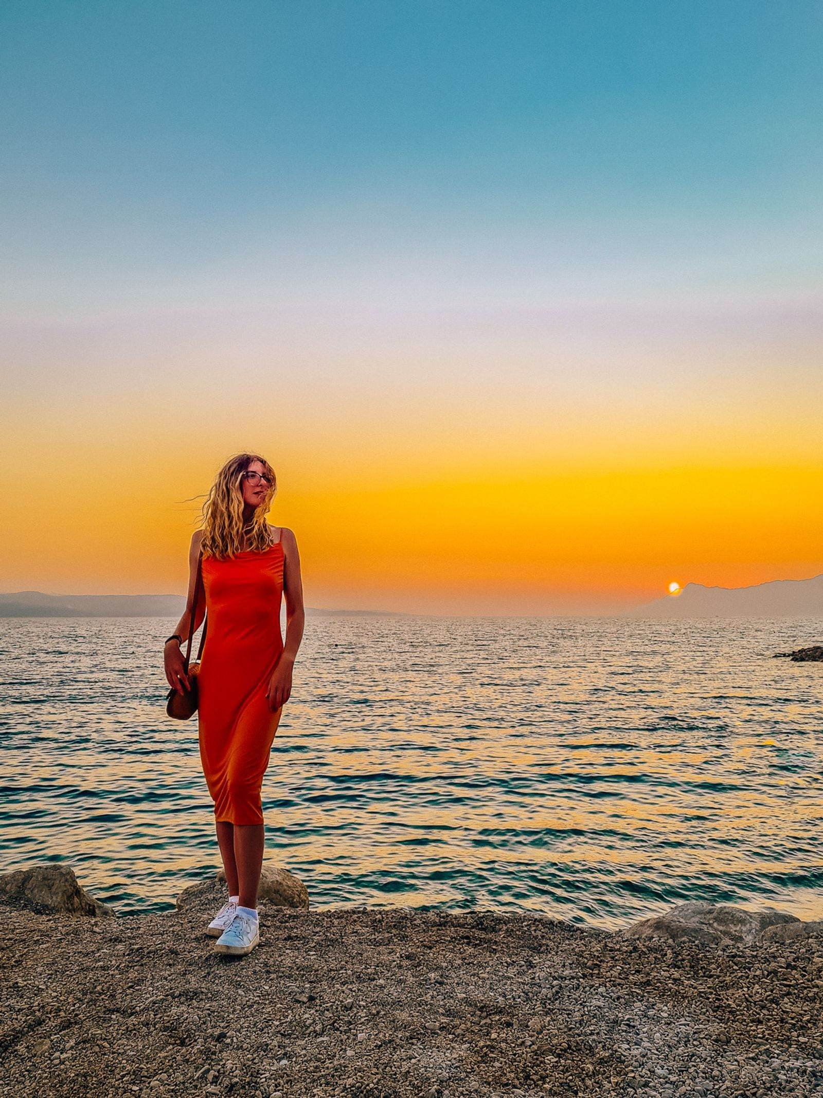 girl in an orange dress standing on the shore with the sea behind and orange sky at sunset