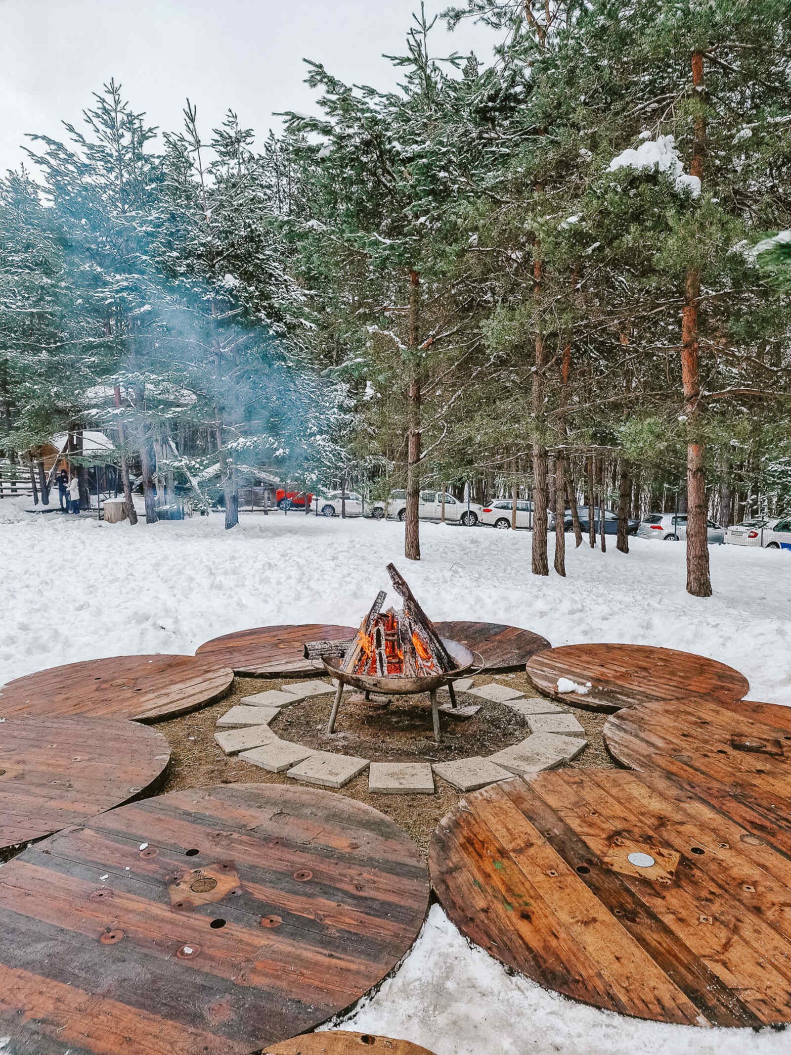 a fire pit with a roaring log fire in the centre of wooden decking with snow on the ground
