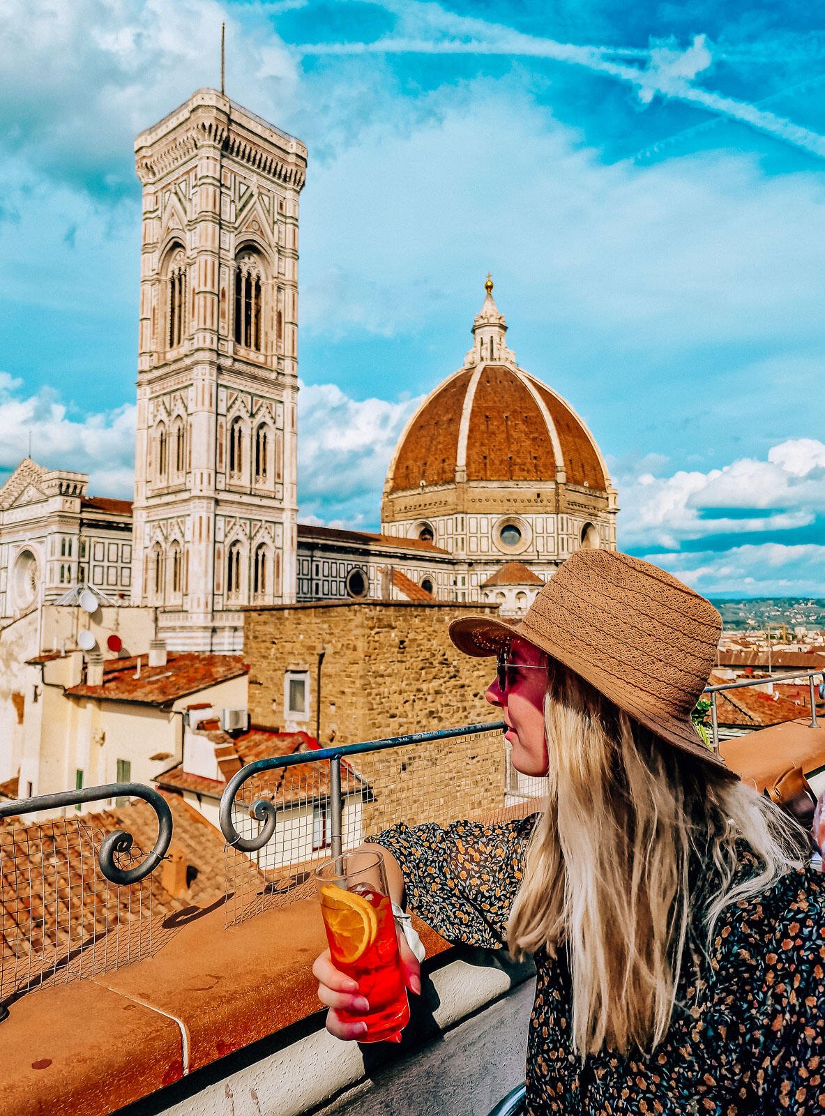 girl in a brown wide rim hat, holding an orange drink looking out across rooftops to the view of the Florence Duomo