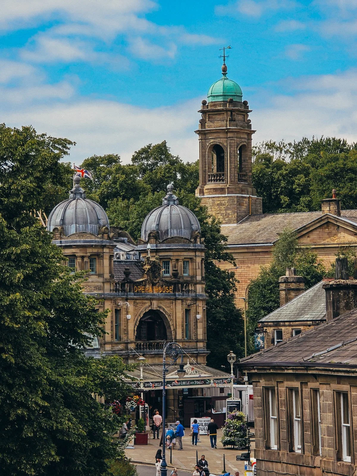 Buxton opera house with people standing outside, the building is detailed, made of sandstone with an old-dashioned awning, narrow glass winsows and two domed turrets