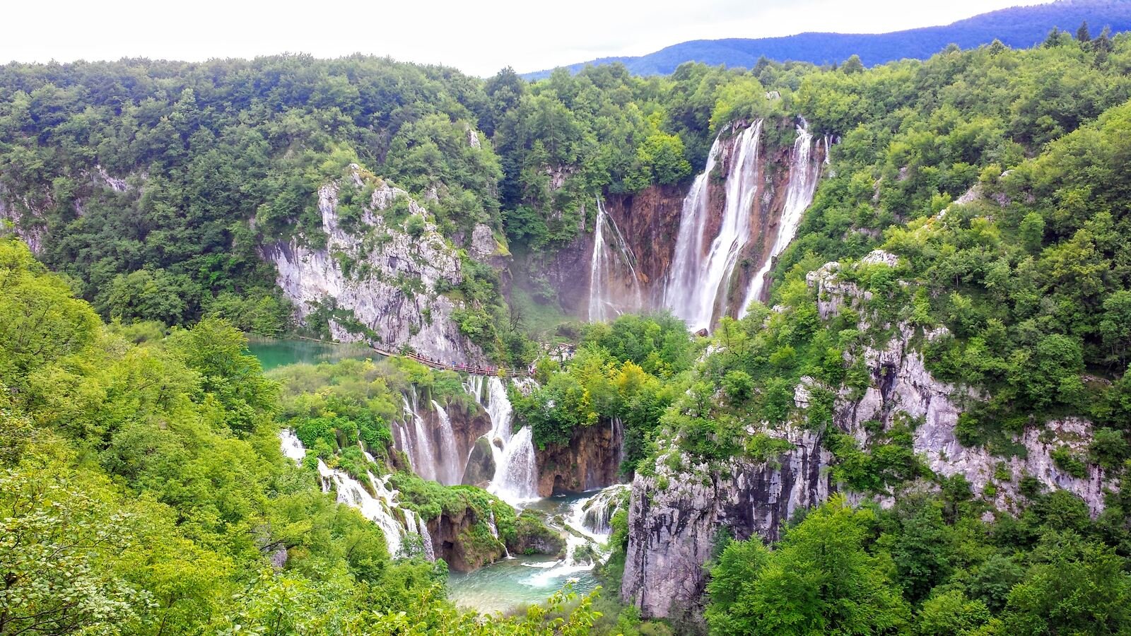 A landscape view of waterfalls tumbling down cliffsides into turquoise pools at the bottom in Plitvice National Park, Croatia. The water is surrounded by lush greenery