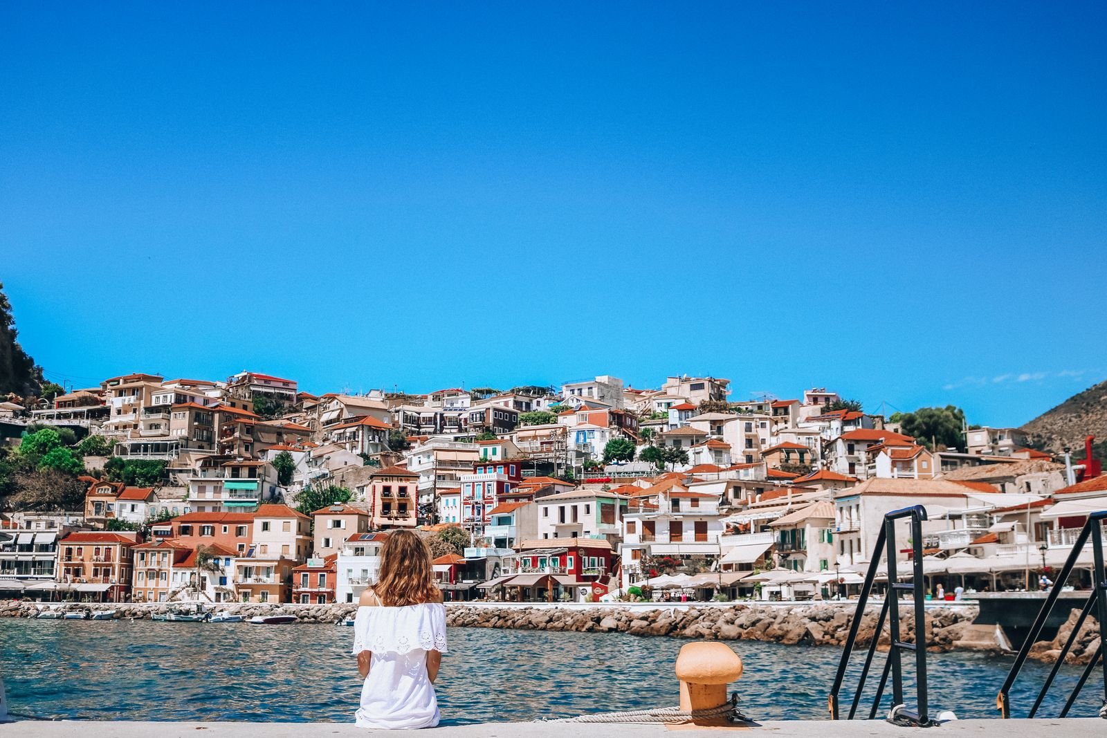 Girl in white dress sitting on a dock looking at Parga town hillside
