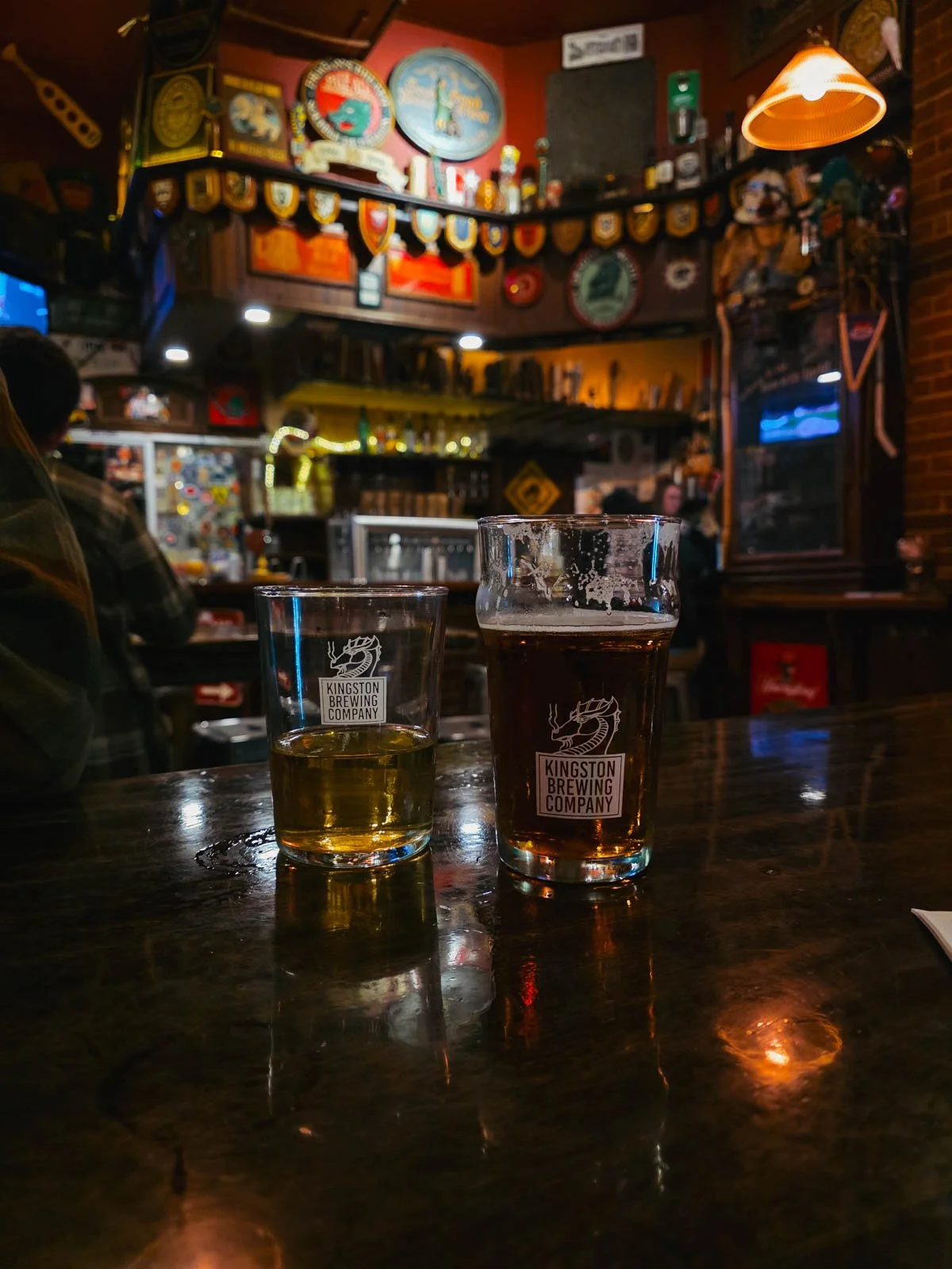 Two glasses of beer on a dark wooden table at a brewery with many colourful decorations along the bar in the background