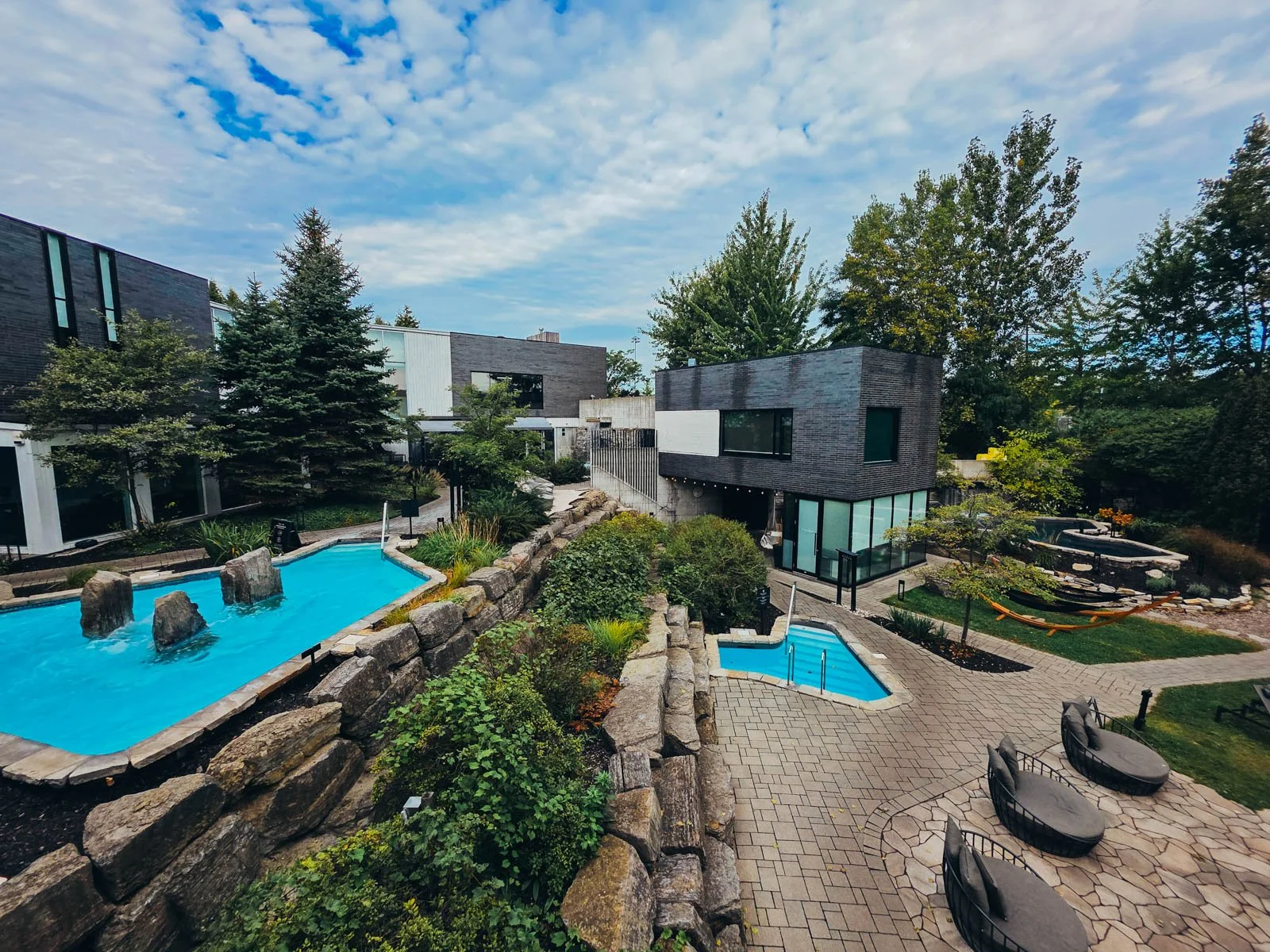 A high view of the large outdoor area of a spa with three blue pools, modern grey buildings, adirondack chairs, and modern buildings all surrounded by green shrubs