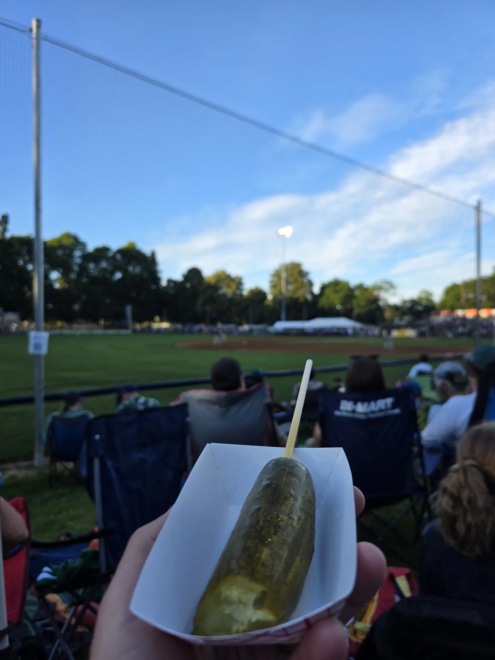 A pickle on a stick in a white cardboard pickle tray at a baseball game