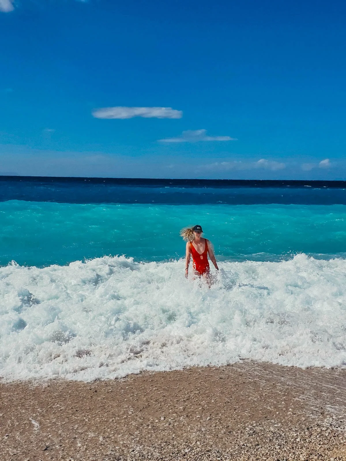 Helena in a red swimsuit at one of the best beaches in the Albanian Riviera, standing in the middle of whit crashing waves with clear blue water behind her