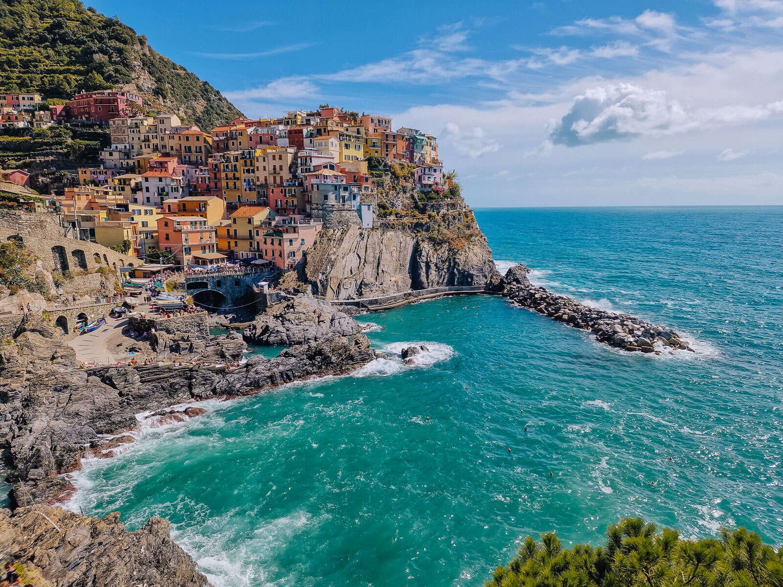 Panoramic view point of Manarola looking across the harbour with turquoise water and rocky shoreline with the colourful houses of Manarola Cinque Terre on the cliff, in orange, pink, yellow. Green hills to the left and sea out to the right. Blue sky