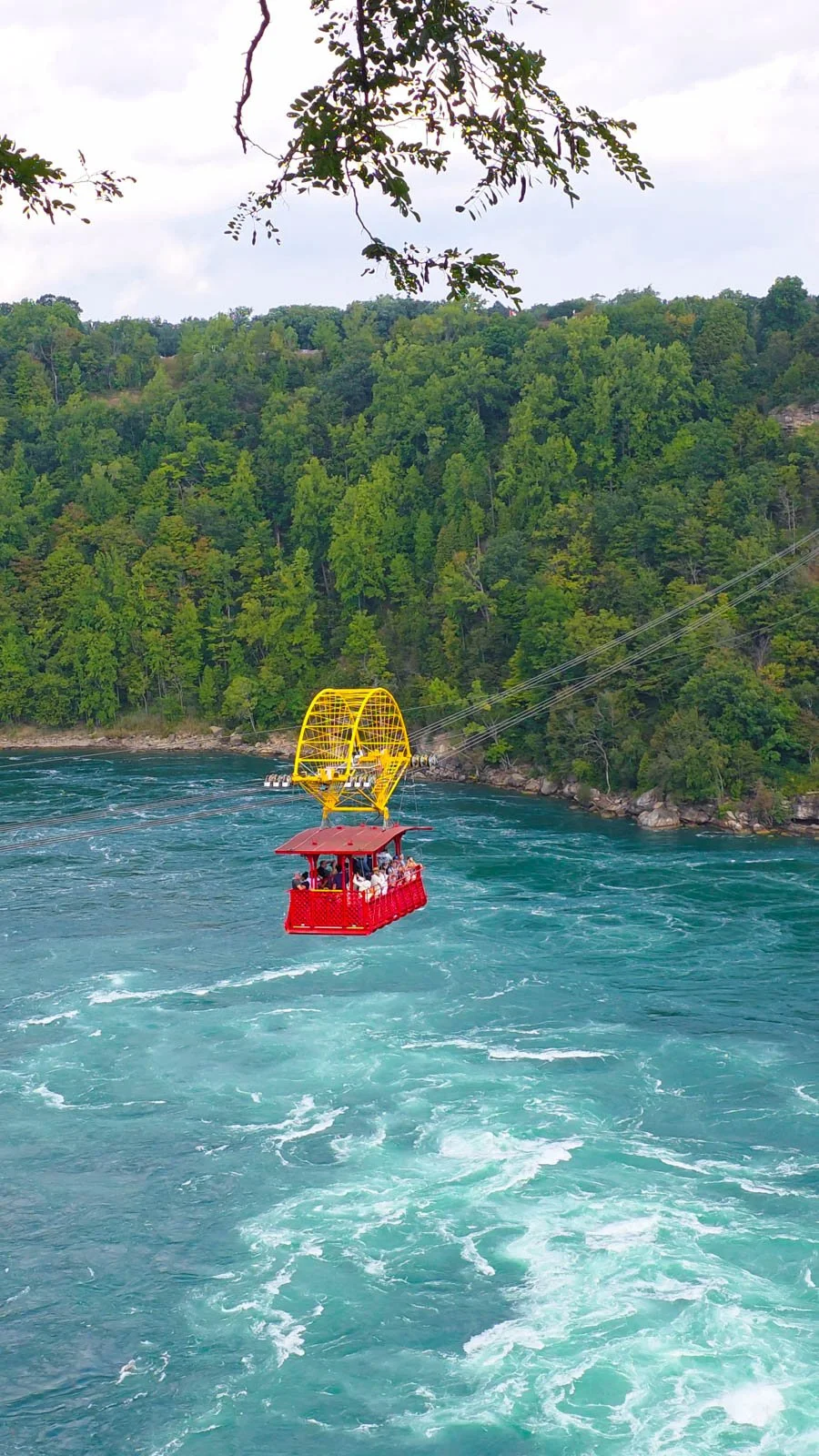 A red metal cable car with a yellow wheel on the cables, suspended over a turquoise pool of water with white crests on the fast-flowing water. Green trees are on the banks of the river