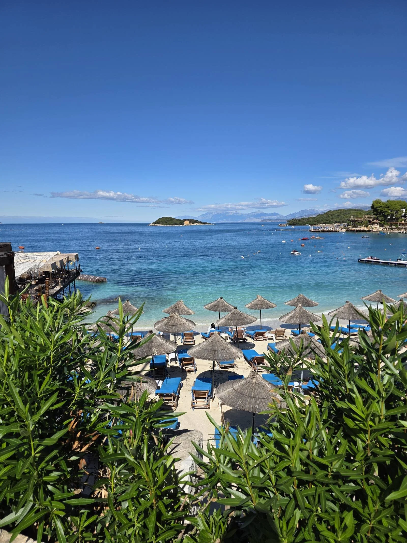 Looking through a gap in greenery at a white sand beach with many parasols and blue sunbeds with calm, turquoise sea beyond on a clear, sunny day