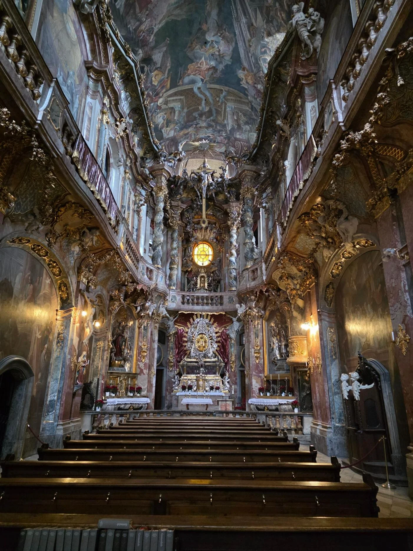 A the interior of a inredablble ornate church with murals pained all over its walls, ornate fixtures covered in gold, and many rows a dark wooden church pews