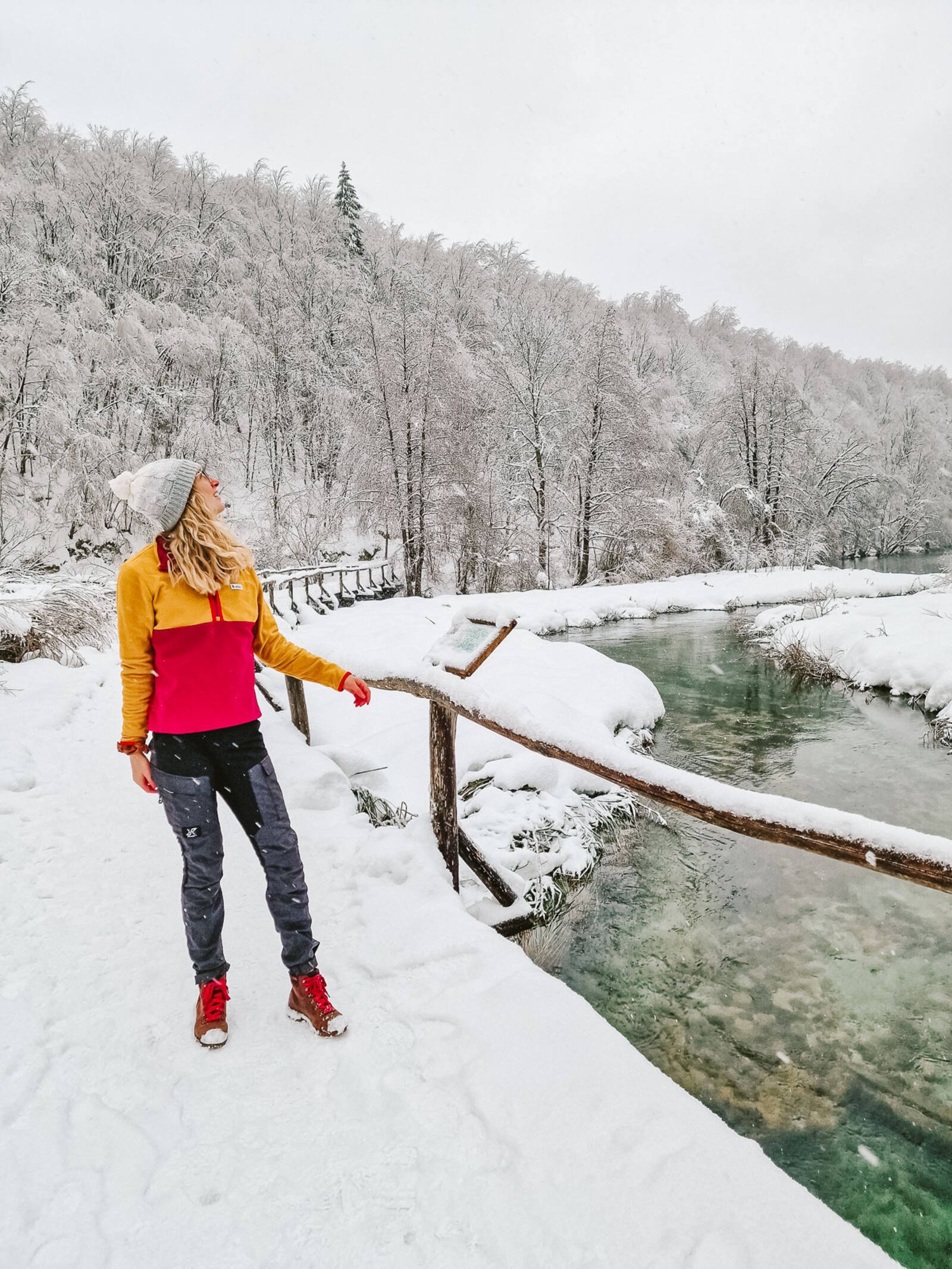 Woman standing on a snow covered bridge with a river running underneath and snow covered trees on the hill in the background. Woman is dresses in black trousers and a bright pink and yellow sweater and hat - packing list Plitvice National Park winter