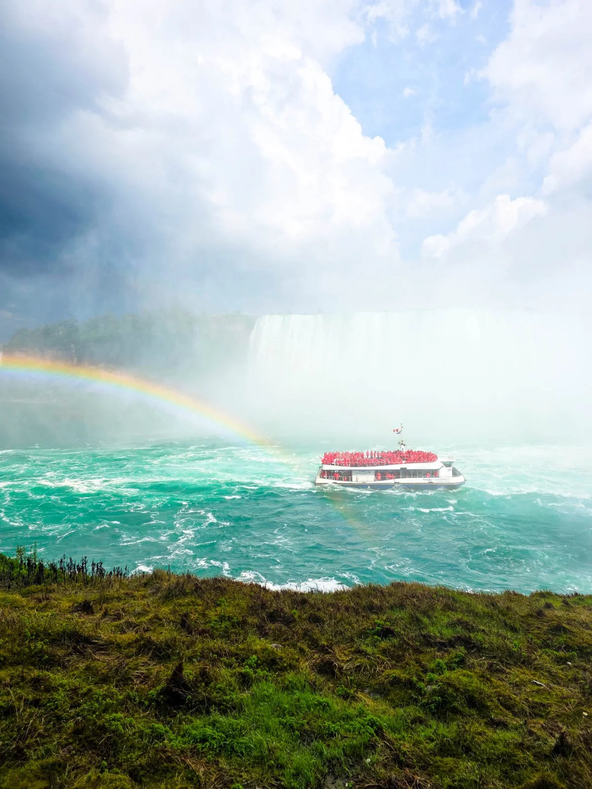Looking down from a hill o a full boat with people in red ponchos cruising blue water with a waterfall and rainbow behind the boat