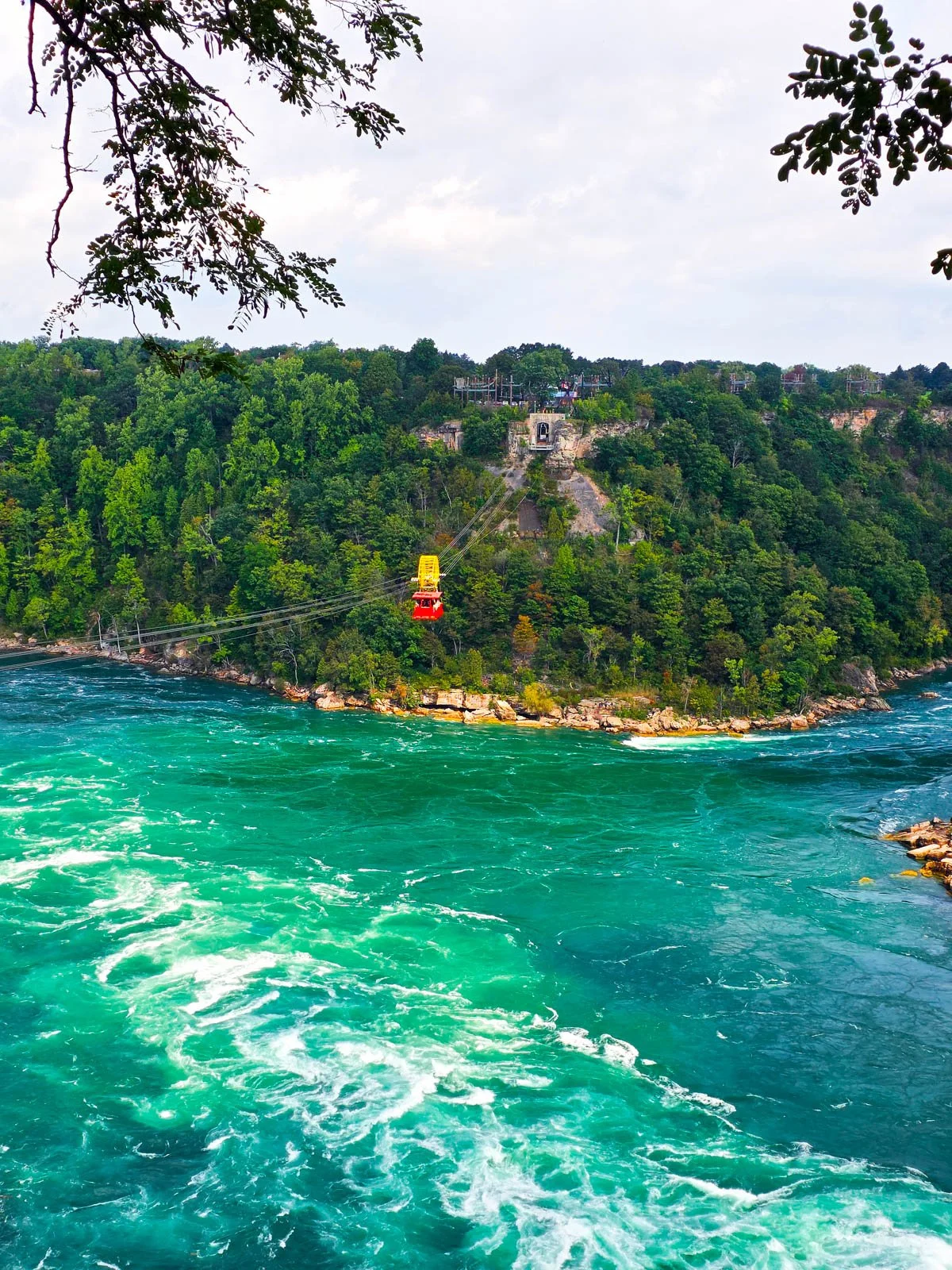 A red metal cable car with a yellow wheel on the cables, suspended over a turquoise pool of water with white crests on the fast-flowing water. Green trees are on the banks of the river