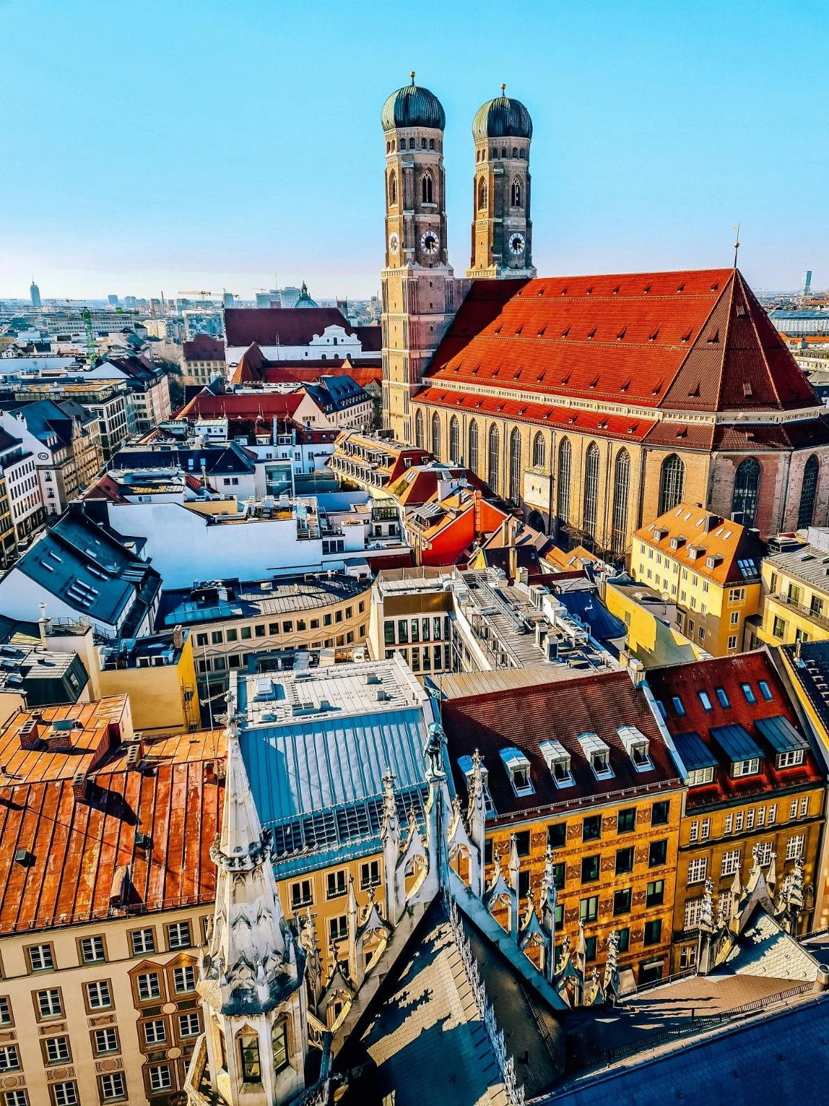 A roof top view of a large church with a red roof and two large domed clock faced towers. The chuch is surrounded by many old colourful buildings in Munich's old town