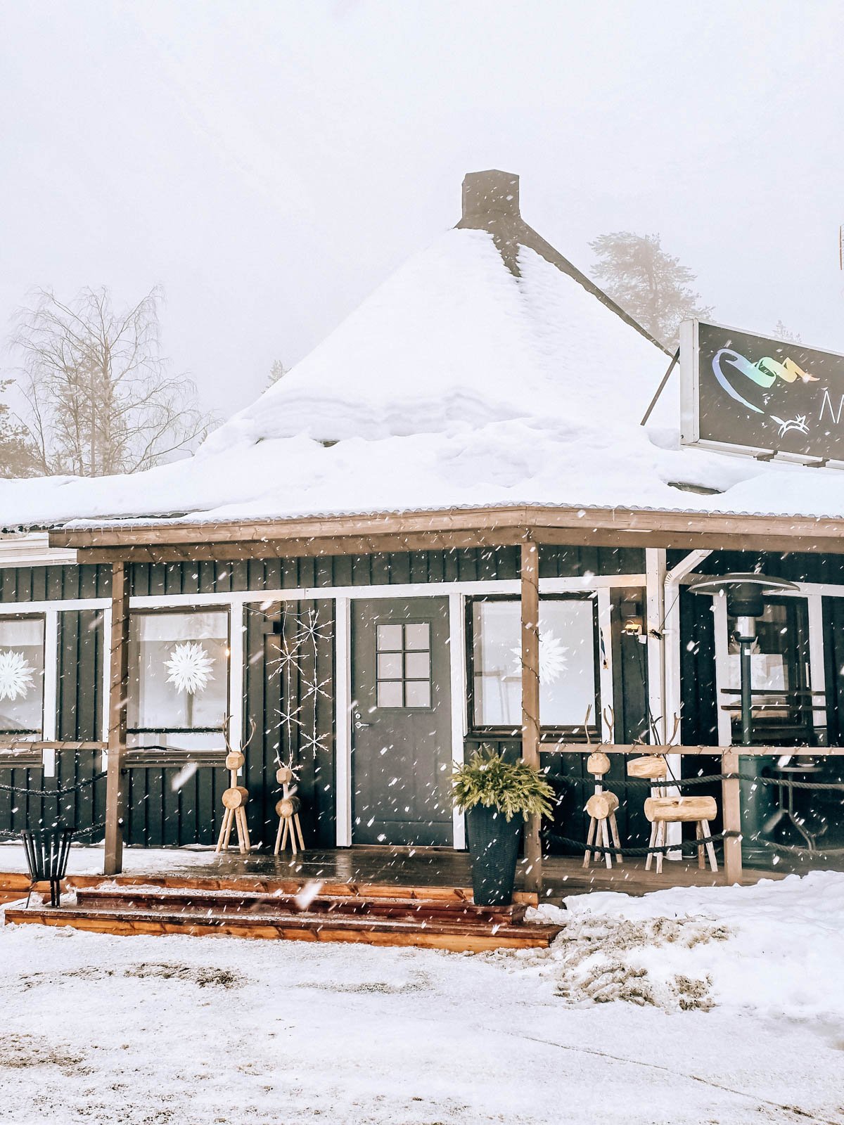 A large dark wooden snow covered building with a pointed roof and large deck. Four wooden reindeer are out on the deck