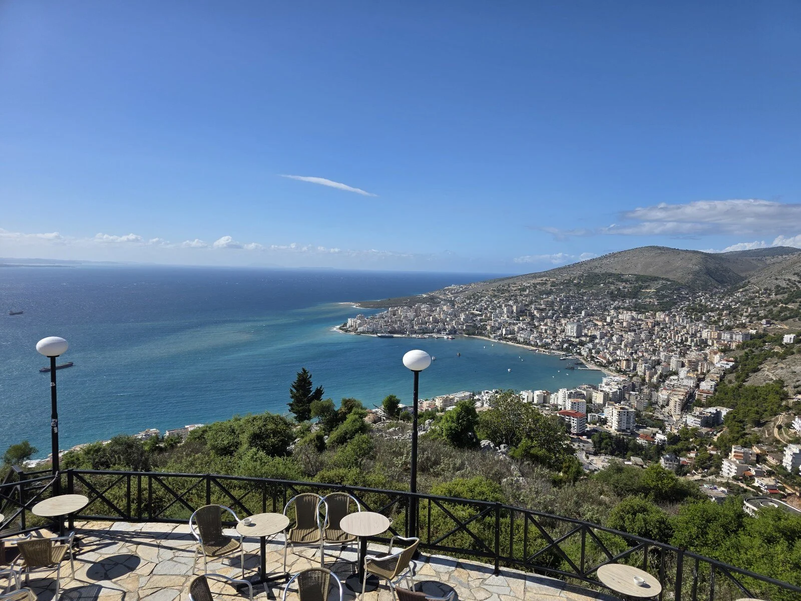 a viewing platform with outdoor seating looking down on a bay with blue water and blue sky. A town is built around the bay