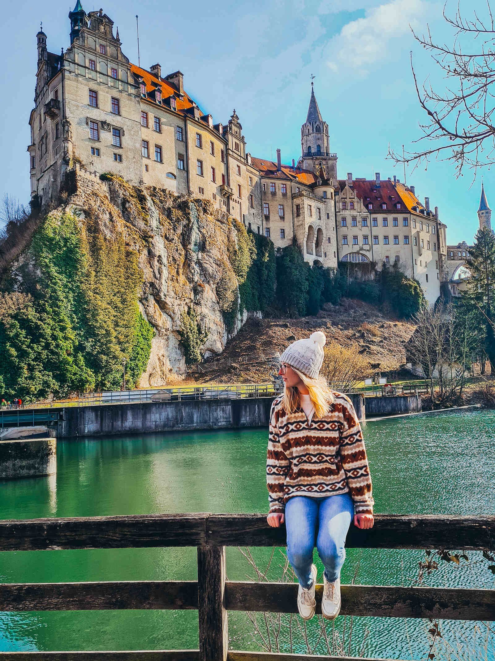 Girl sitting on a fence that lines a river and across the river is a large castle that sits on top of a rocky cliff