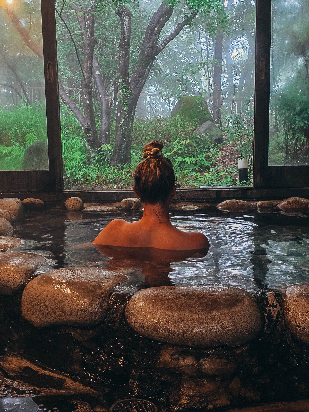 A woman in a small stone heated onsen pool looking out of open wooden doors into the wooded area beyond