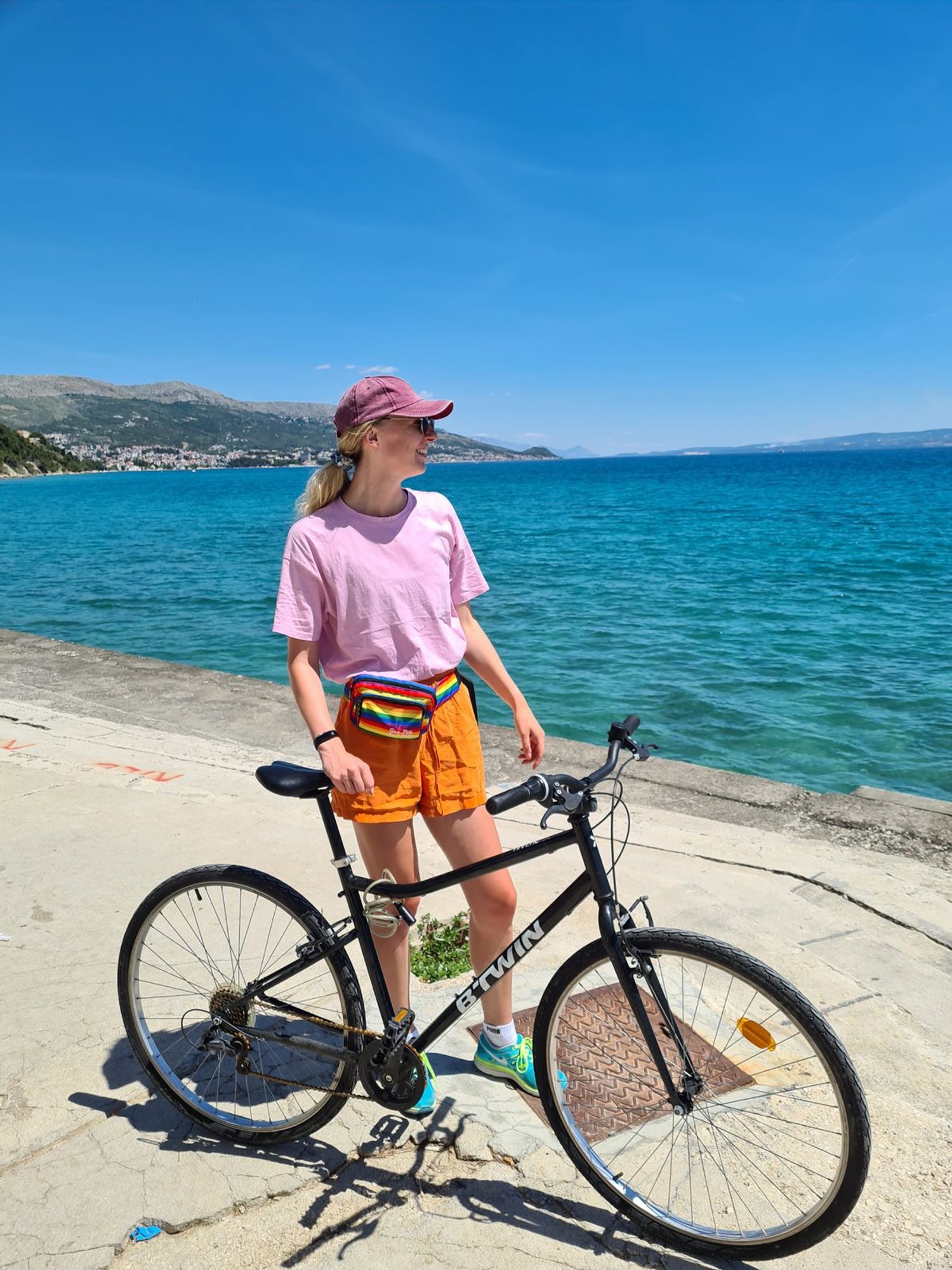 girl in pink tshirt and orange top standing next a bike alongside the sea