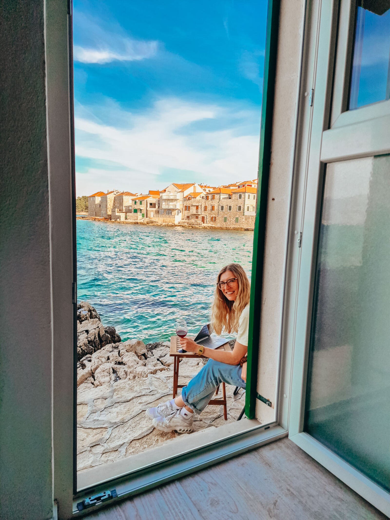 girl sitting at a table with a laptop on it outside a door that's right next to the sea. She's holding a glass of wine and stone houses are in the background
