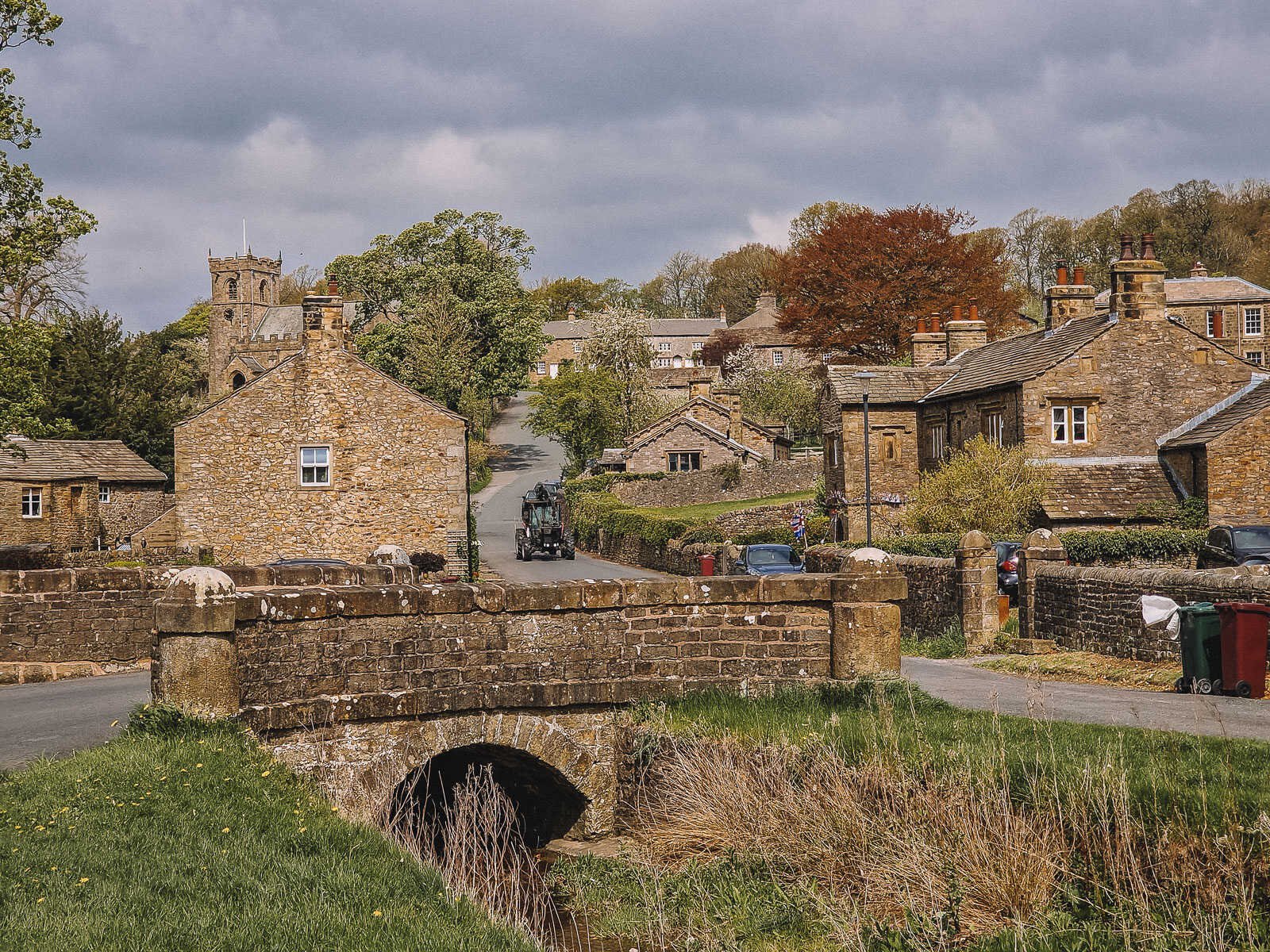 a charming village with sandstone coloured cottages and a small bridge crossing a stream. A tractor approaching down the hill in the distance