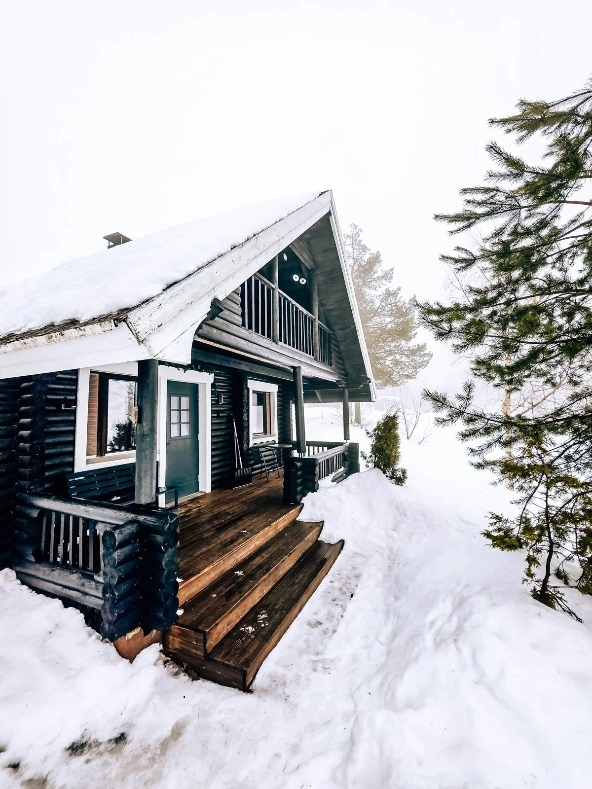 A dark coloured wooden cabin with a porch and steps leading up to it, surrounded by snow at Aava Sky Village accommodation in Aavasaksa