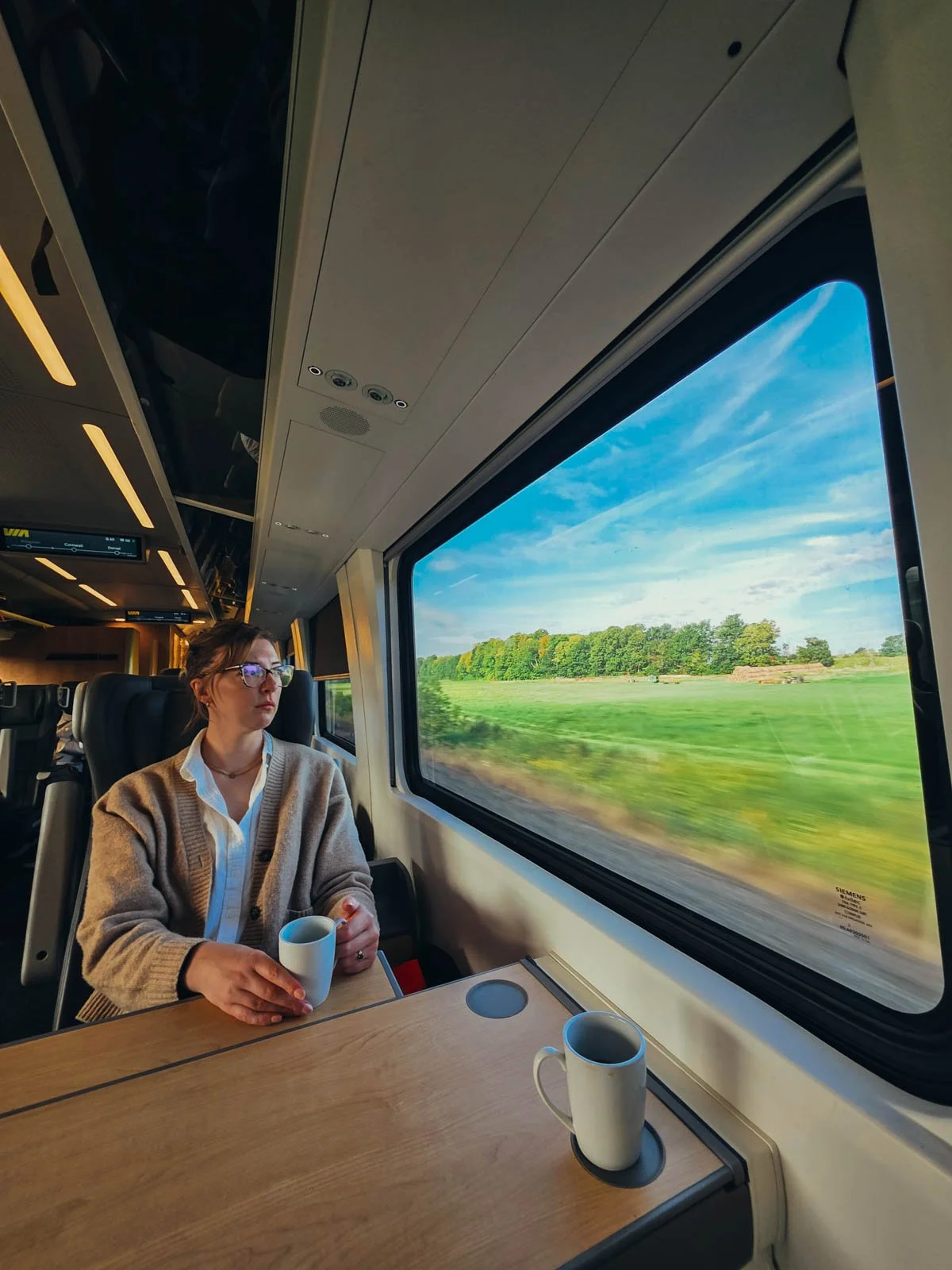 Helena sitting at a train table in VIA Rail Business Class in Canada, holding a cup of coffee and looking out the window at a green landscape with trees and blue sky