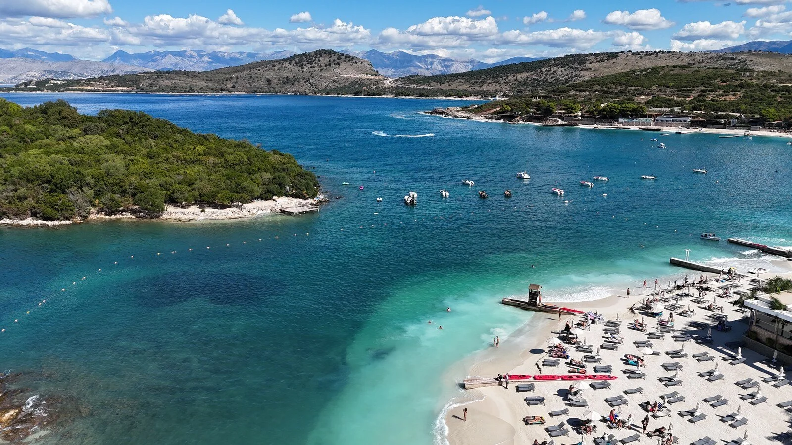 a drone shot of blue and turquoise sea with a white sand bech in the right corner, many islands and coastline spread out beyond with lots of boats in the water on a sunny day