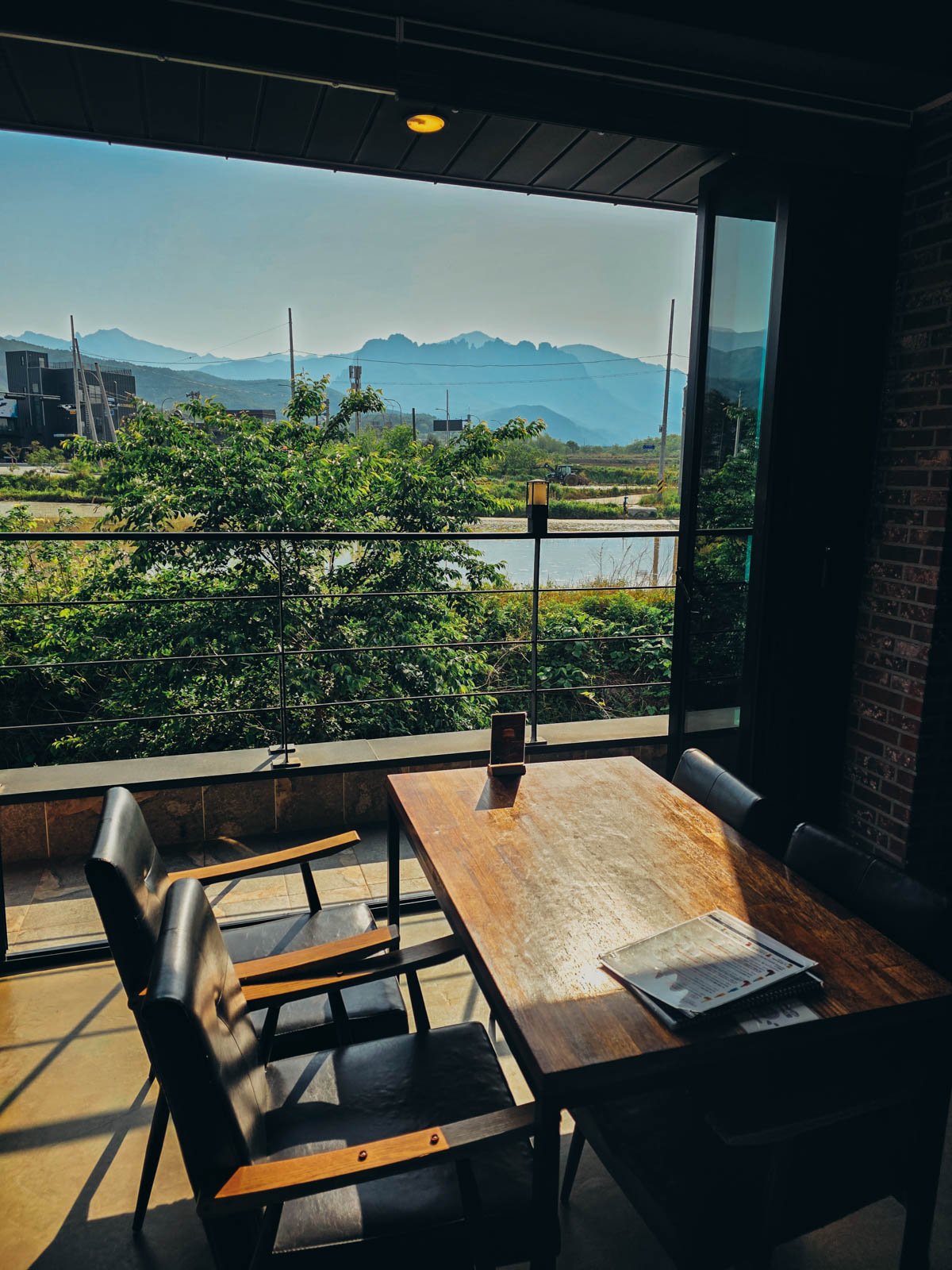 A table in a brewery sitting at a wide open window looking out over a balcony and rice paddies to a hazy mountain range beyond
