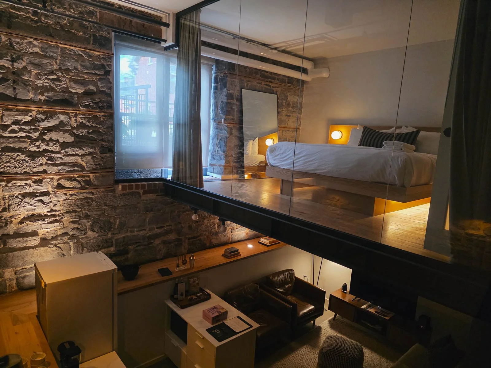 A large hotel room in a loft style with a bedroom on the second floor seen through a glass wall above the hotels seating area and kitchen which can be seen below