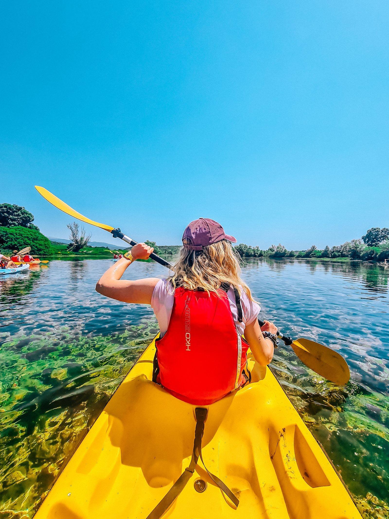 back of a girl wearing a red lifevest and sitting in a yellow kayak with both hands on a paddle, paddling down a rive rin the sun