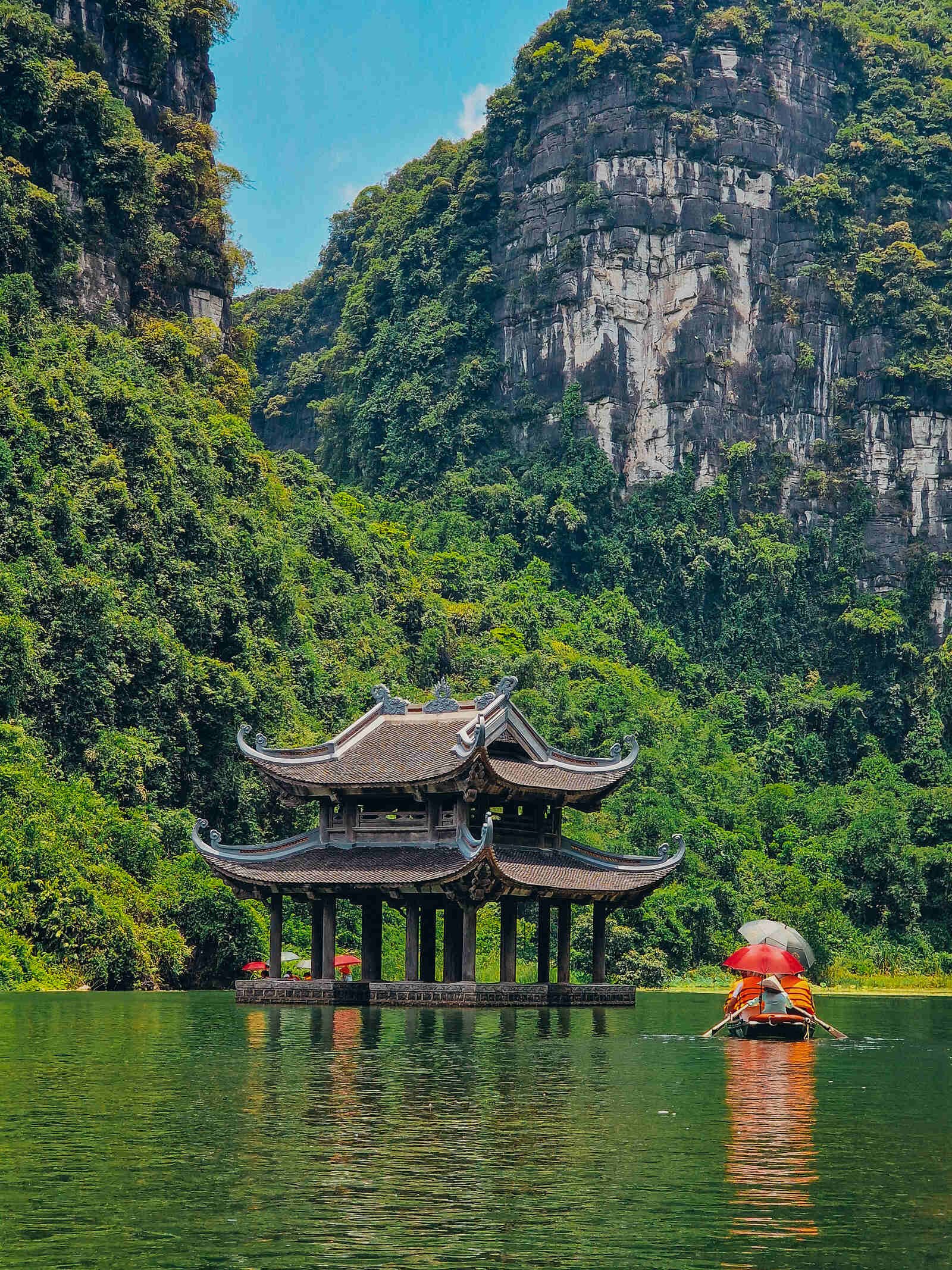 a wooden pagoda in the middle of a waterway surrounded by grren cliff faces. A small boat with red umbrellas in it rows past the pagoda