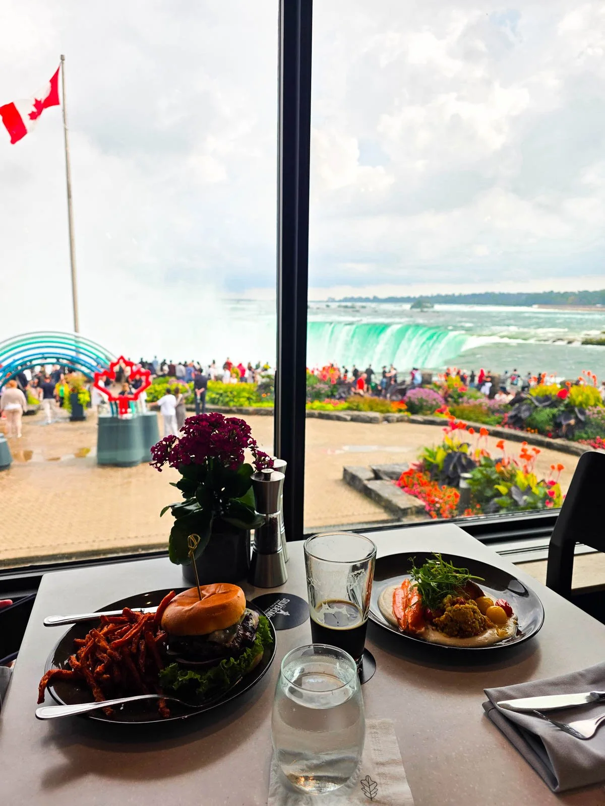 Two plates of food on the table of a restaurant next to a window with a view of Niagara Falls and a Canadian glaf flying outside