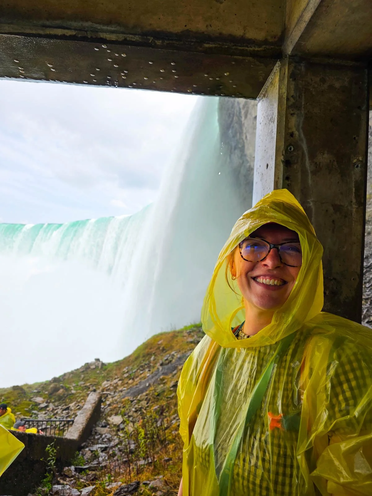 Helena in a yellow poncho with the hood up smiling at the camera and the powerful water flowing over Niagara Falls horseshoe falls behind her