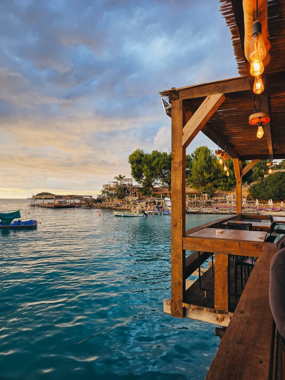 The wooden decking of a beach bar along clear blue water and another beach side bar in the distance