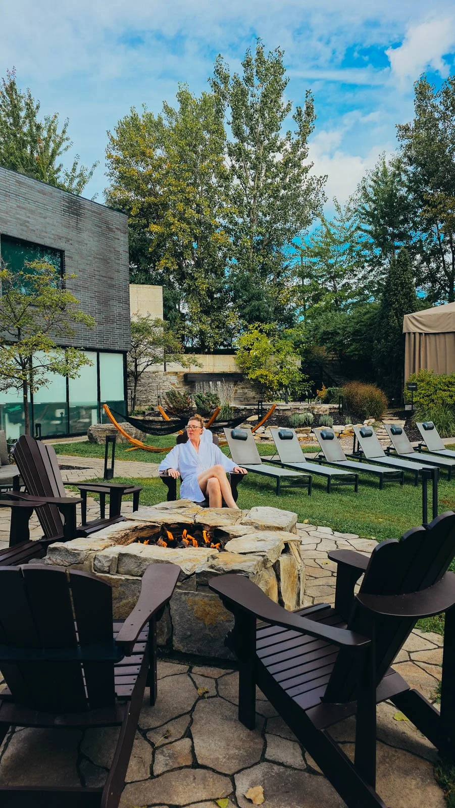 Helena in white sitting outside in front of a stone fire pit with adirondack chairs around it and spa buildings behind her